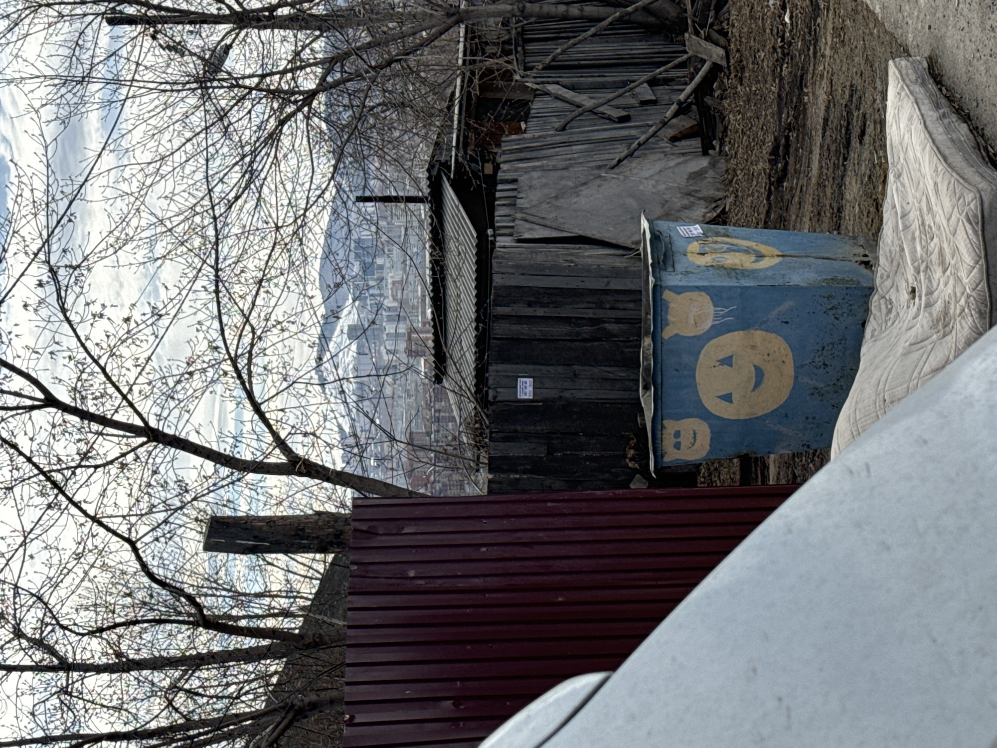 A slightly rotated outdoor shot focuses on a cluttered scene. A blue rectangular bin with yellow smiley-face graffiti sits beside a discarded mattress. Behind them, a dilapidated wooden structure stands near a maroon corrugated fence. A tree with bare branches reaches across the scene, with buildings visible in the distant background. This image was taken in the city of Krasnoyarsk, Russia.