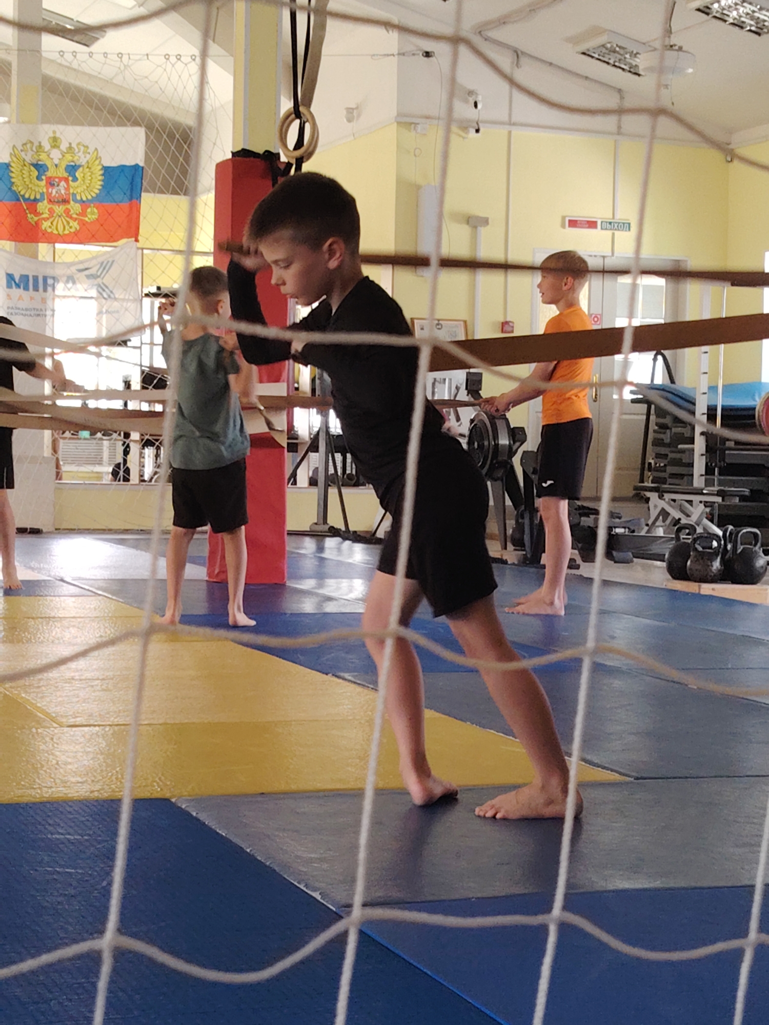 The image, taken indoors in what appears to be a gymnasium in the city of Chaikovsky, Russia, depicts a group of young boys engaged in physical activities. A Russian flag is visible in the background. One boy in the foreground, wearing a black shirt and shorts, stands barefoot on a blue and yellow mat. To his left, another boy in a green shirt and black shorts is also barefoot. To his right, a third boy in an orange shirt and black shorts stands near a rowing machine, also barefoot. Weights are visible in the background. The foreground is obscured by what appears to be a net.