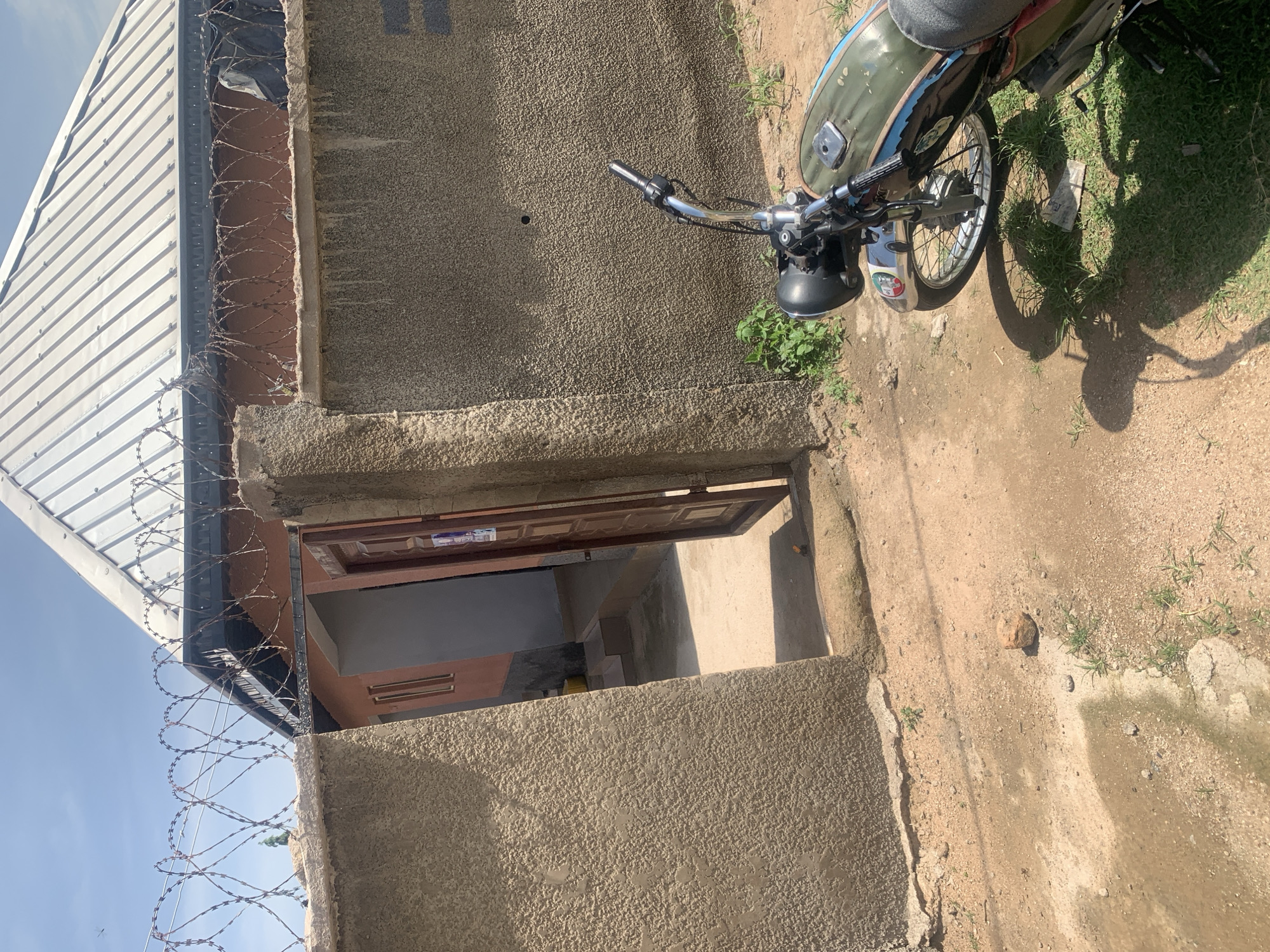 The image captures a scene outside a building in Jauro Yinu, Nigeria. A motorcycle, partially visible on the right, is parked on a dirt and grass surface. The motorcycle's handlebars and headlight are prominent, with its green fuel tank showing some wear. Green grass and dry earth make up the ground.
The building in the background features a rough, textured concrete exterior. A section of a corrugated metal roof is visible on the left. Barbed wire is strung along the top of the wall, suggesting a need for security. An open doorway leads into the interior, revealing a dark hallway and what appears to be wooden doors. The lighting suggests it is daytime, likely with clear skies. The overall impression is one of a utilitarian and somewhat austere environment. There is no discernible text in the image.