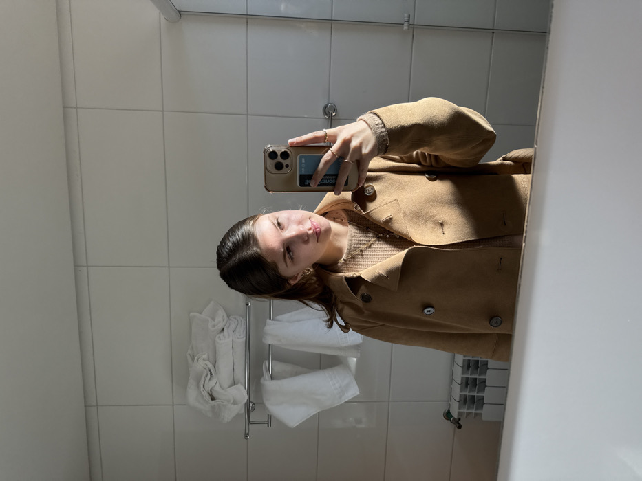A female subject is depicted in a bathroom, identified as being located in the city of Canela, Brazil, taking a mirror selfie. The subject, visible from the chest up, has light brown hair and is wearing a tan double-breasted coat over a light brown knitted sweater. She accessorizes with gold hoop earrings, a pendant necklace, and multiple rings on her right hand. She holds a gold-colored smartphone with a clear case in her right hand, with a blue card visible within the case. The background consists of white tiled walls with a chrome towel rack on the left holding folded and draped white towels. Below the towel rack, a white heating radiator is visible. The foreground features the white reflective surface of a bathtub rim or counter, with part of a glass shower enclosure visible on the far right. Natural light from the right casts shadows across the subject and the tiled wall.