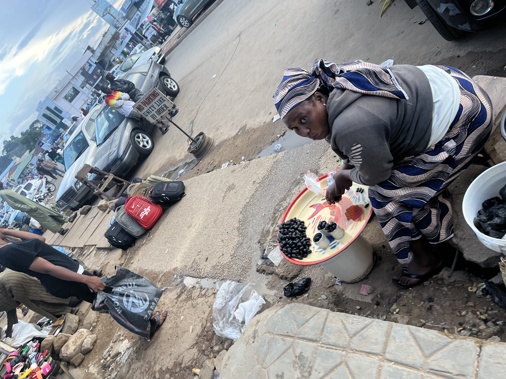 The image depicts a bustling outdoor market scene, likely in Jos, Nigeria. In the foreground, a woman wearing a headscarf and a patterned skirt is attending to her wares. She is crouched over a shallow white bucket and a red and yellow tray filled with dark, round fruits, possibly olives or dates. Next to the tray, there are small metal cups also containing some of these fruits. Her facial expression suggests focus on her task.
Behind the woman, several people are visible, some appearing to be shoppers and others vendors. A person in a black shirt and striped pants is holding a black plastic bag with visible text. Bags of various colors, including red and grey, are stacked on the ground near parked cars. One car, a silver sedan, is prominent in the middle ground, with a "NO PARKING UNITY LINE BY ORDER" sign attached to a pole beside it. The ground is a mix of concrete, dirt, and some puddles, indicating a potentially wet or recently rained-on environment.
In the background, more vehicles and market stalls are visible, suggesting a lively commercial area. The sky is partly cloudy, hinting at a daytime scene. The overall impression is one of everyday commerce in a busy urban or semi-urban setting in Nigeria.