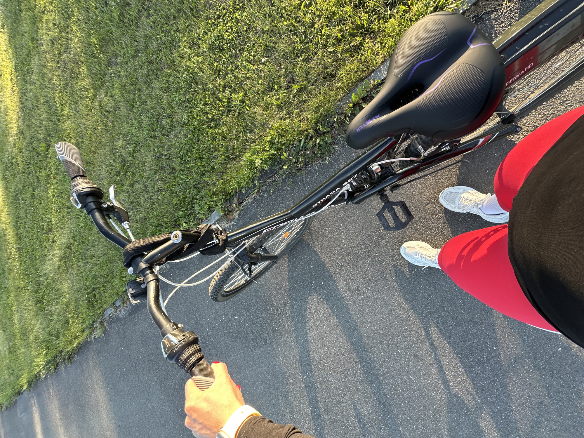 This image shows a person's perspective from behind the handlebars of a bicycle, facing forward. The bicycle is black with some white and red accents, and has a black seat with purple detailing. The bicycle is on an asphalt path next to a grassy area. The person is wearing red leggings and white athletic shoes. Their left hand is visible, holding the left handlebar grip, and their right hand is out of frame. The sun is casting long shadows, suggesting it is either early morning or late afternoon. The scene is outdoors, likely in a park or on a bike trail. There is no discernible text on the bicycle or clothing. The overall impression is one of a recreational activity, possibly a bike ride in pleasant weather.