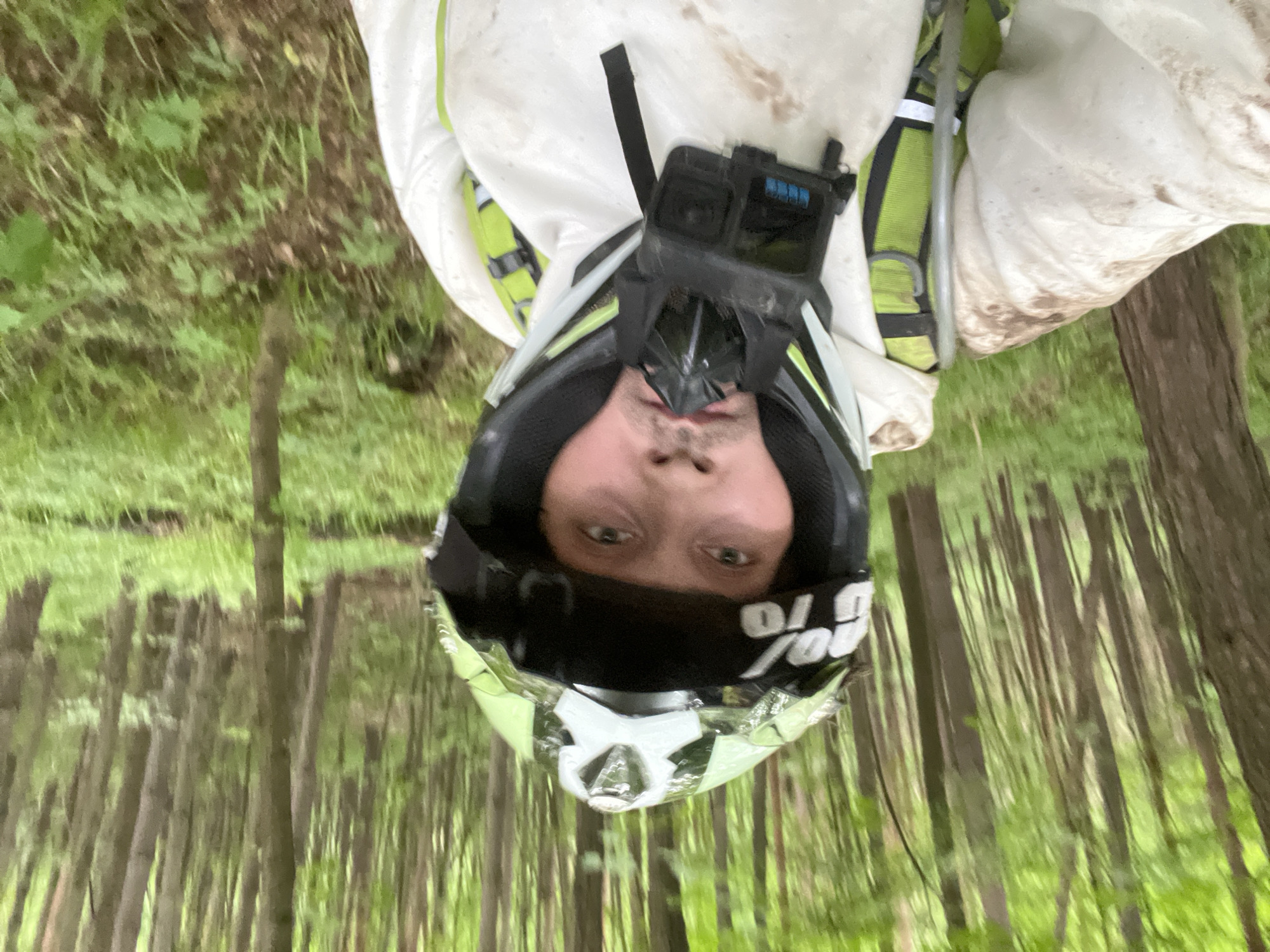 A man in a dirt bike helmet and white protective suit stands in a forest. The helmet has a GoPro attached to the top. The background shows closely packed trees and green vegetation, suggesting a wooded area. The man is facing the camera directly. The trees resemble the kind of forest that grows in Russia. The location of Gruzinskii, Russia, cannot be determined from the image.