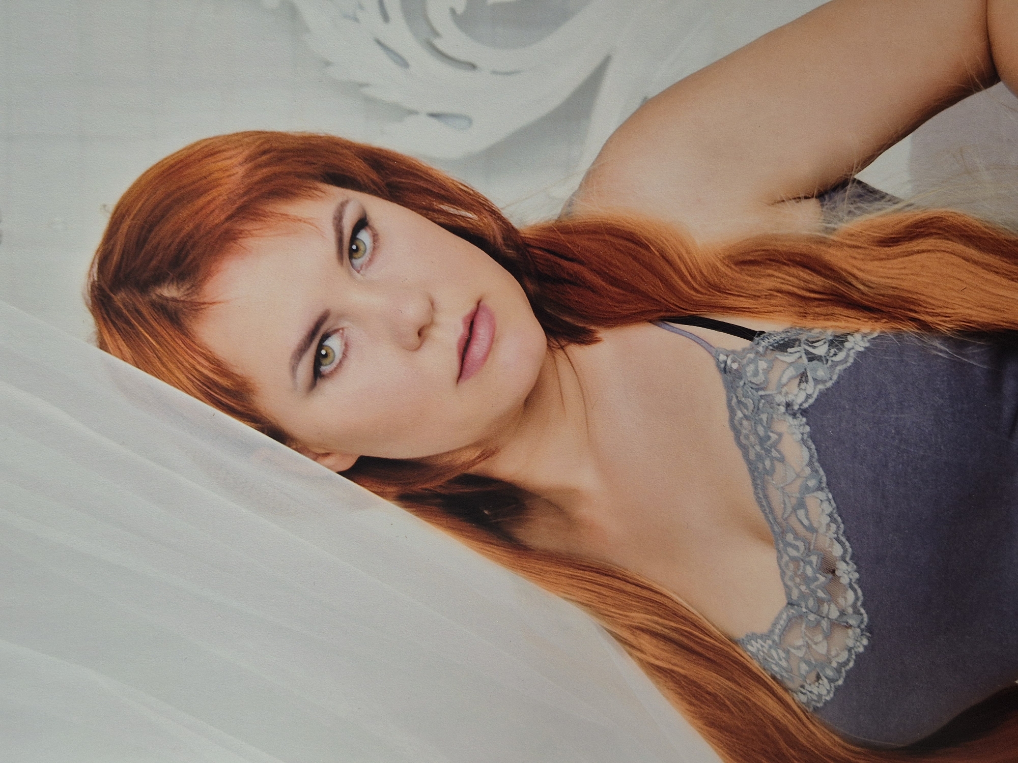 The image is a medium shot portrait of a young woman with long, wavy auburn hair. She is wearing a dark grey tank top with a delicate lace trim. Her gaze is directed slightly off-camera to her left, and her lips are parted. She is positioned in front of a light-colored backdrop with a subtle, abstract pattern. The woman's face is illuminated, and she appears to be indoors. The image does not contain any elements that would suggest a specific location within Schwäbisch Gmünd, Germany, or any other geographical area.