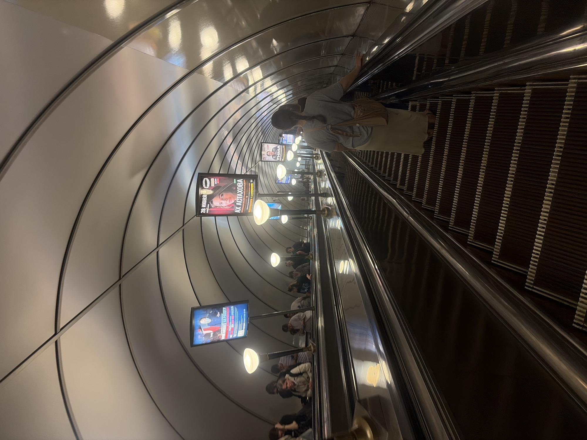 This image captures a detailed view of a deep underground metro escalator hall in Saint Petersburg, Russia, specifically at Admiralteyskaya station.
The scene features three parallel escalators ascending towards the station exit/platform level. The perspective is from one of the ascending escalators, looking forward and slightly upwards. In the immediate foreground, a person with dark hair in a braid, wearing a light grey short-sleeved top, a long cream-colored skirt, and carrying a light brown over-the-shoulder bag, stands on the rightmost escalator, facing away from the camera with their left hand resting on the silver handrail.
The escalators themselves have dark brown, ridged steps with light horizontal stripes, polished silver handrails, and dark, reflective metallic side panels that show subtle reflections of the overhead lights and structure. Further up the escalators, approximately 15-20 other individuals are visible, also ascending. They are casually dressed; some are looking at their mobile phones.
The most prominent architectural feature is the tunnel-like ceiling, which consists of numerous uniformly curved, highly reflective, silver-colored metallic panels. These panels are arranged in parallel, creating a ribbed effect, and are separated by thin, dark horizontal strips. This structure forms a continuous overhead enclosure.
Illumination is provided by multiple large, spherical, white light fixtures mounted on tall, slender, dark poles positioned strategically between the escalator lanes.
Several large, vertical digital display screens are suspended from the ceiling structure. A central screen on the left clearly shows Russian text, including "28 ИЮНЯ" (June 28) and "АХ АСТАХОВА" (Akh Astakhova), accompanied by a portrait of a woman. Further up, another screen displays "20 ИЮЛЯ" (July 20) and "ТИТОМА" (Titoma) with a different person's image. A screen on the far left shows an image resembling a news broadcast with a man in uniform.
The overall color scheme is dominated by metallic silver, various shades of grey, dark brown, and black, contrasted by the bright white light from the fixtures and the illuminated advertisements. The environment appears functional, well-lit, and well-maintained.