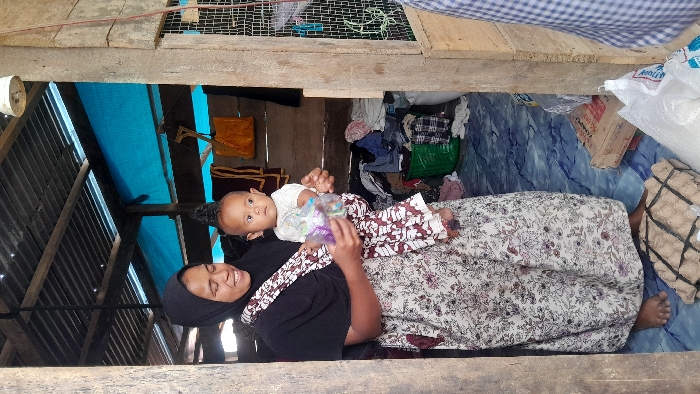 A smiling woman in a black hijab and floral dress lies on her side, holding a baby in her arms. The baby, dressed in a light shirt and patterned pants, looks up and to the left, grasping a plastic bag. They are inside a room with corrugated iron walls and a cluttered space filled with various belongings. The room is sparsely furnished, with a blue sheet covering part of the wall and a patterned mattress.