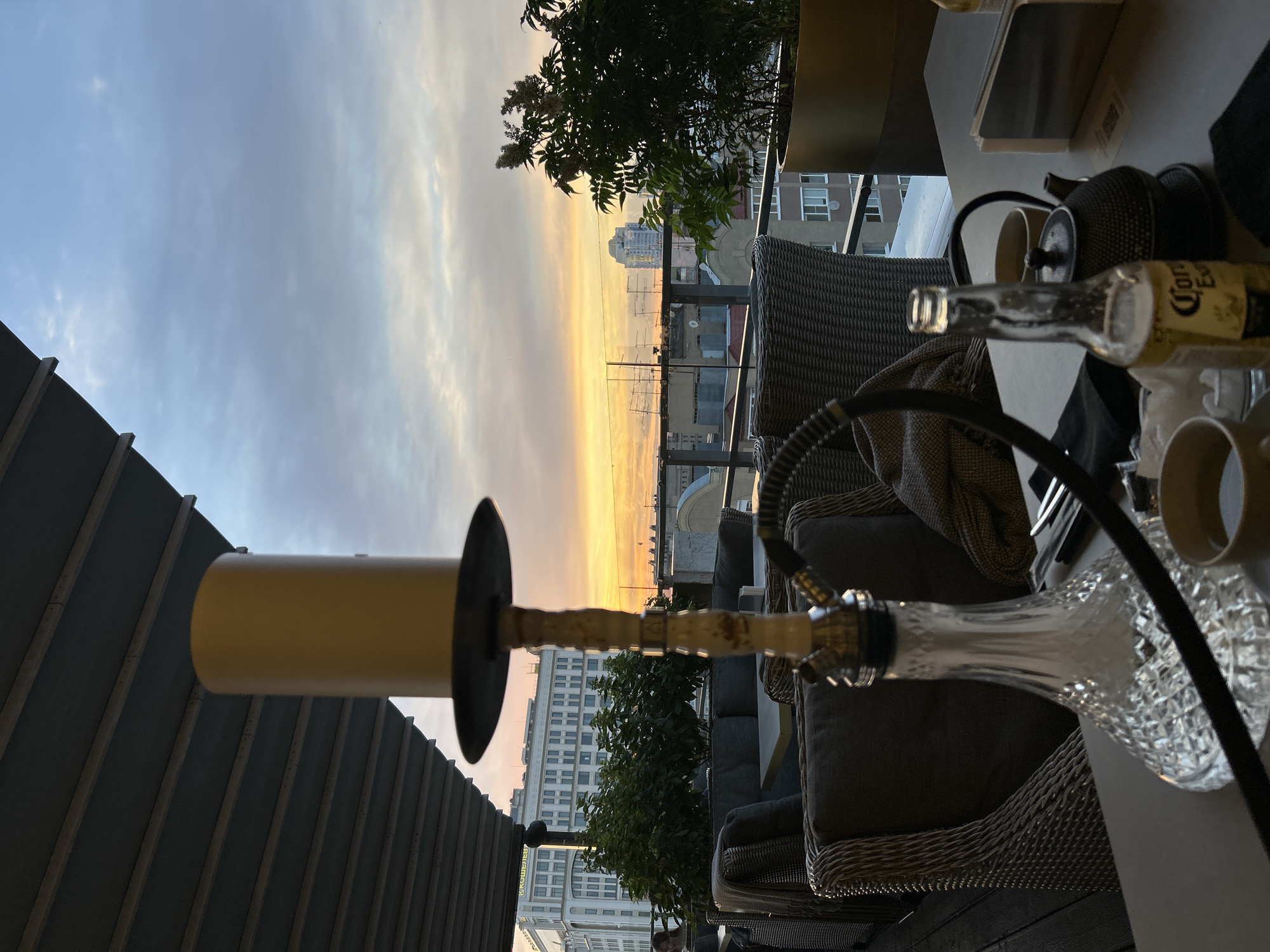 The photograph depicts a rooftop seating area at dusk. A shisha pipe is prominently placed in the foreground, with its glass base catching the ambient light. To the right, a bottle of Corona Extra beer, a teapot, and two cups are arranged on a table, suggesting a leisurely gathering. In the background, the cityscape of Novokuybyshevsk, Russia, unfolds under a sky painted with the warm hues of sunset. The architectural style of the buildings suggests a blend of Soviet-era and contemporary designs. The overall atmosphere is one of relaxation and urban enjoyment.