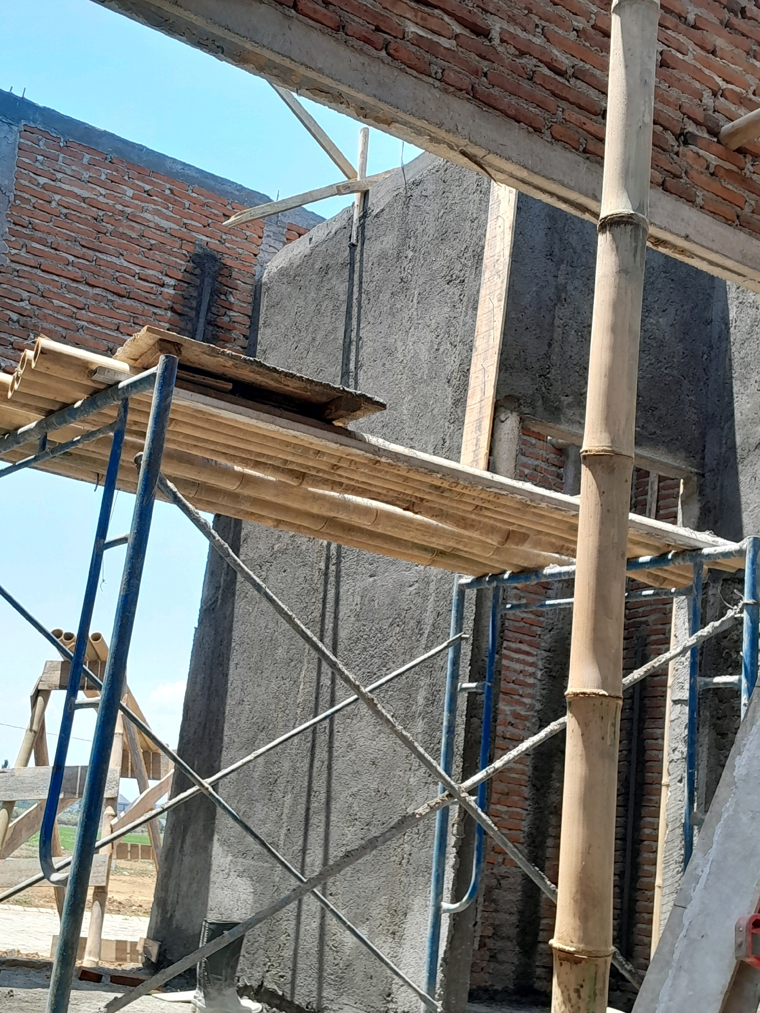 The image depicts a construction site with scaffolding made of bamboo and metal poles supporting bamboo planks. The scene is set outdoors under a clear blue sky, likely in Pakisaji, Indonesia, given the context. A partially constructed building with exposed brickwork and concrete walls dominates the background. A single green boot is visible near the bottom right corner of the image, suggesting the presence of a worker. The overall impression is one of ongoing construction work, with materials and temporary structures arranged for use.