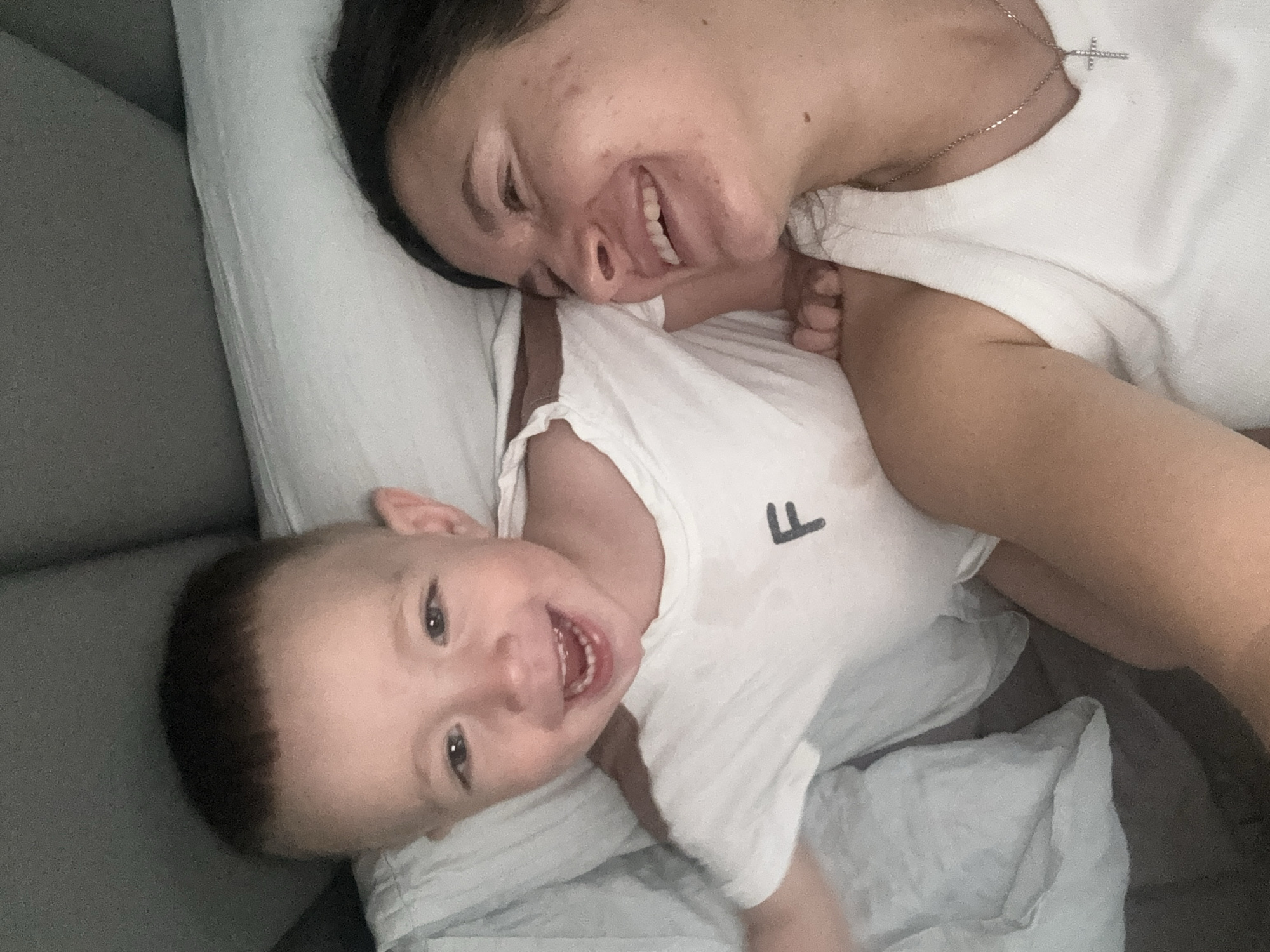 A woman and a baby are lying on a bed, presumably indoors. The woman has dark hair and is wearing a white shirt and a necklace with a cross pendant. She is smiling. The baby, who appears to be a toddler, is also wearing a white shirt with a letter "F" on it and is smiling with their mouth open. They are lying on a white pillow. The background includes a gray couch or bed frame. The location cannot be ascertained from the image but is given as Odintsovo, Russia.