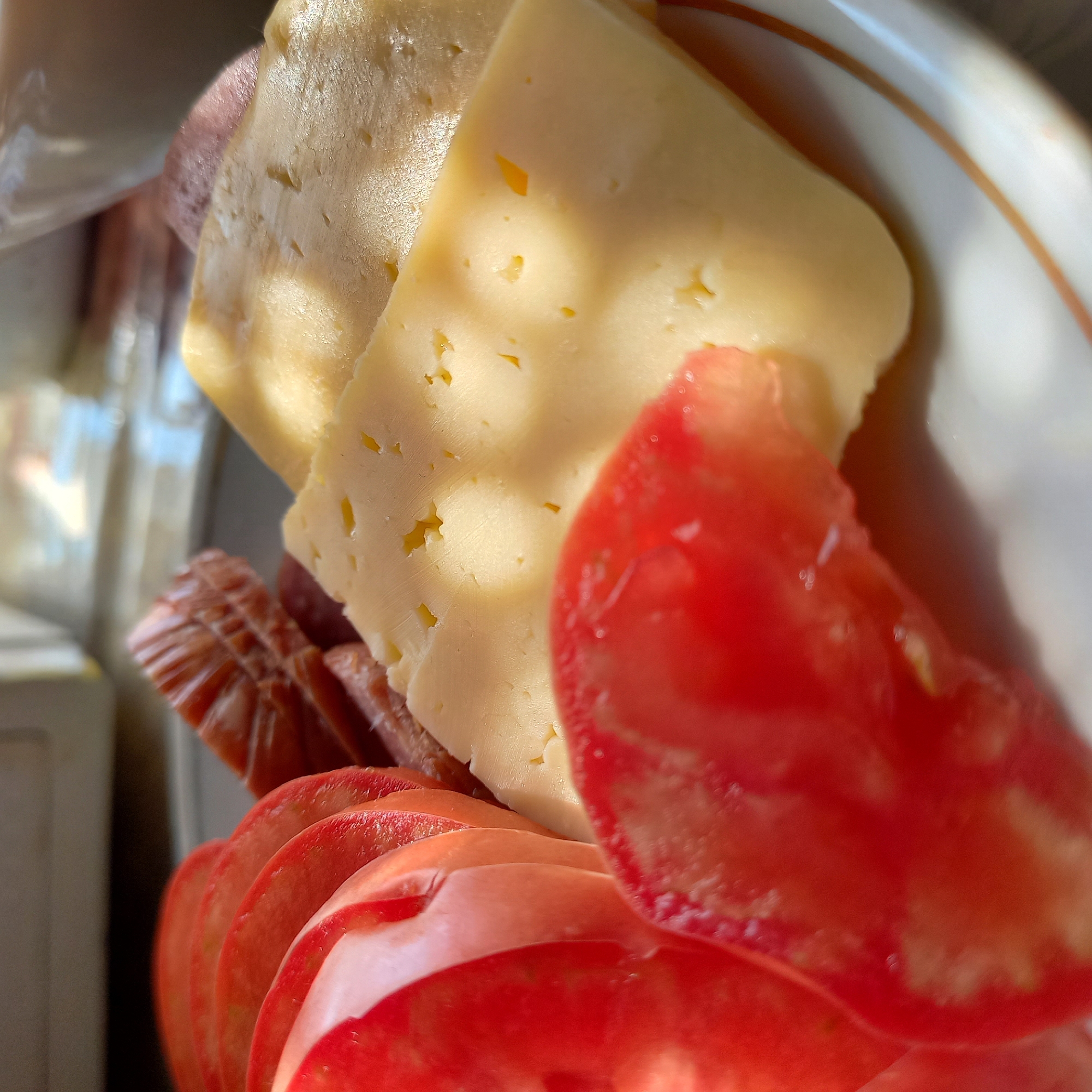 A close-up shot shows a plate of sliced tomatoes, cheese, and a piece of sausage. The tomatoes are bright red and thinly sliced, arranged in a fanned pattern. Two slices of yellow cheese, with small holes, are positioned next to the tomatoes. Behind the cheese and tomatoes, a piece of cured sausage is visible, with its characteristic ridged texture. The food is placed on a white plate with a decorative brown and orange rim. The lighting suggests natural light, with shadows and highlights falling across the food, indicating it might be near a window. The background is blurred, but appears to be an indoor setting, possibly a kitchen or dining area. There are no people visible in the image. The image is a still life of food items.