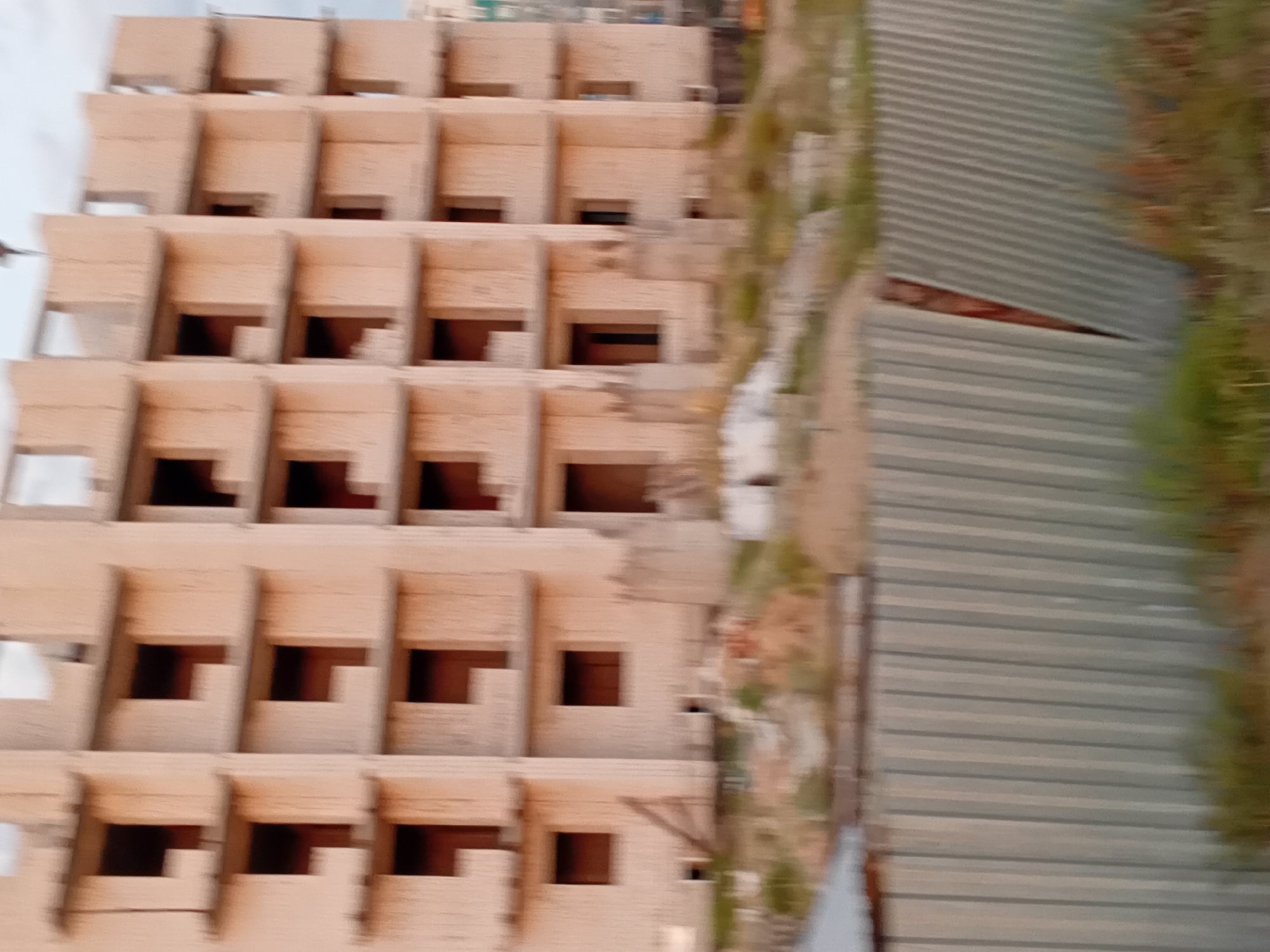 The image shows a multi-story building under construction in the city of Semey, Kazakhstan. The building appears to be made of concrete blocks and has numerous open window frames. The foreground features a corrugated metal fence, partially obscured by vegetation. In the background, other structures and piles of construction materials are visible. The sky is overcast. The overall scene depicts a construction site, likely in an urban or semi-urban environment.