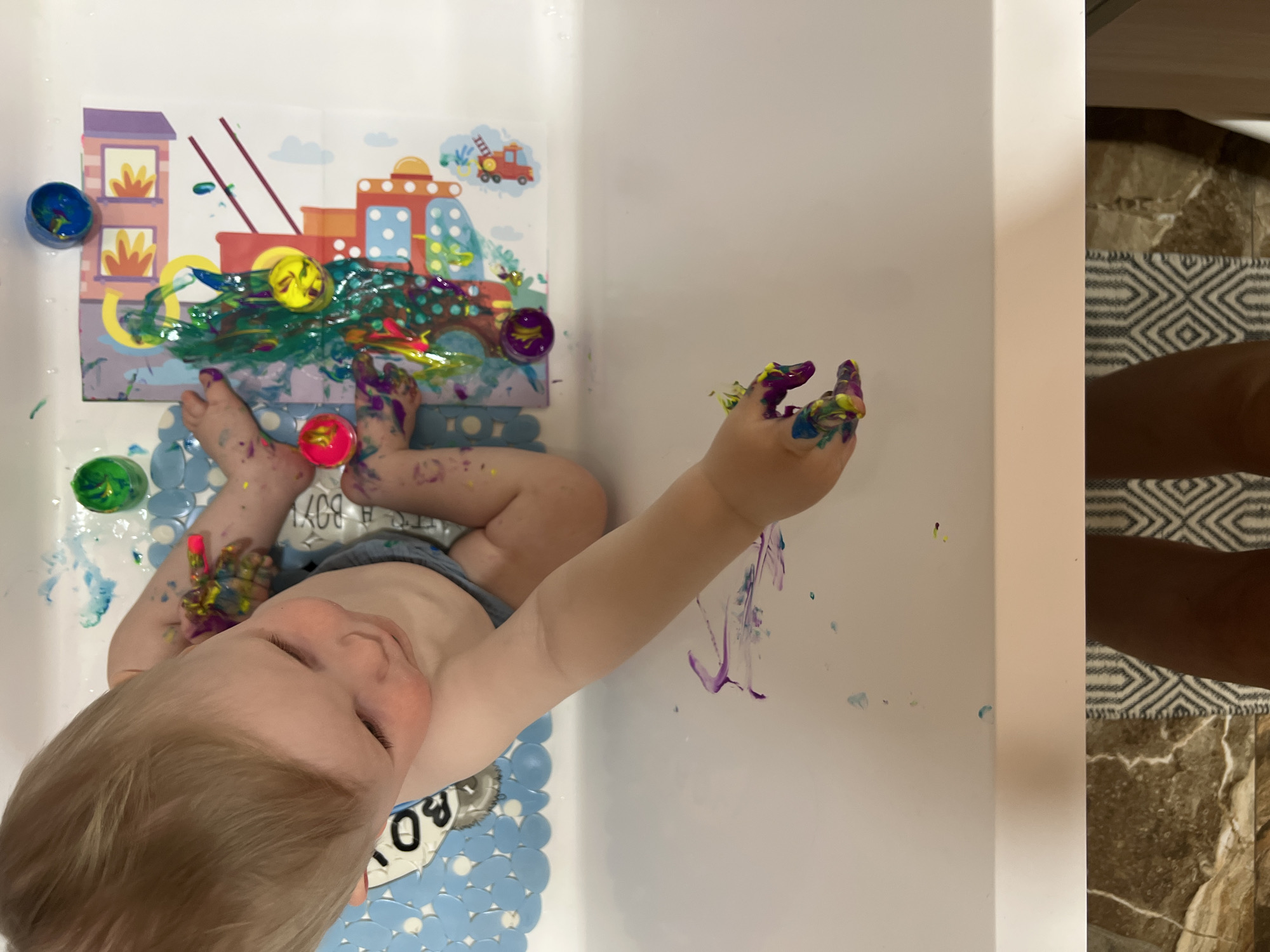 A child sits in a white bathtub. The child has blonde hair, fair skin, and is wearing a diaper. The child's hands and feet are covered in colorful paint. Paint is smeared on the wall and a picture in the bathtub. Paint containers are near the picture. The bottom of a person's legs are seen next to a decorative rug and tile floor in the background. The location cannot be determined from the image.