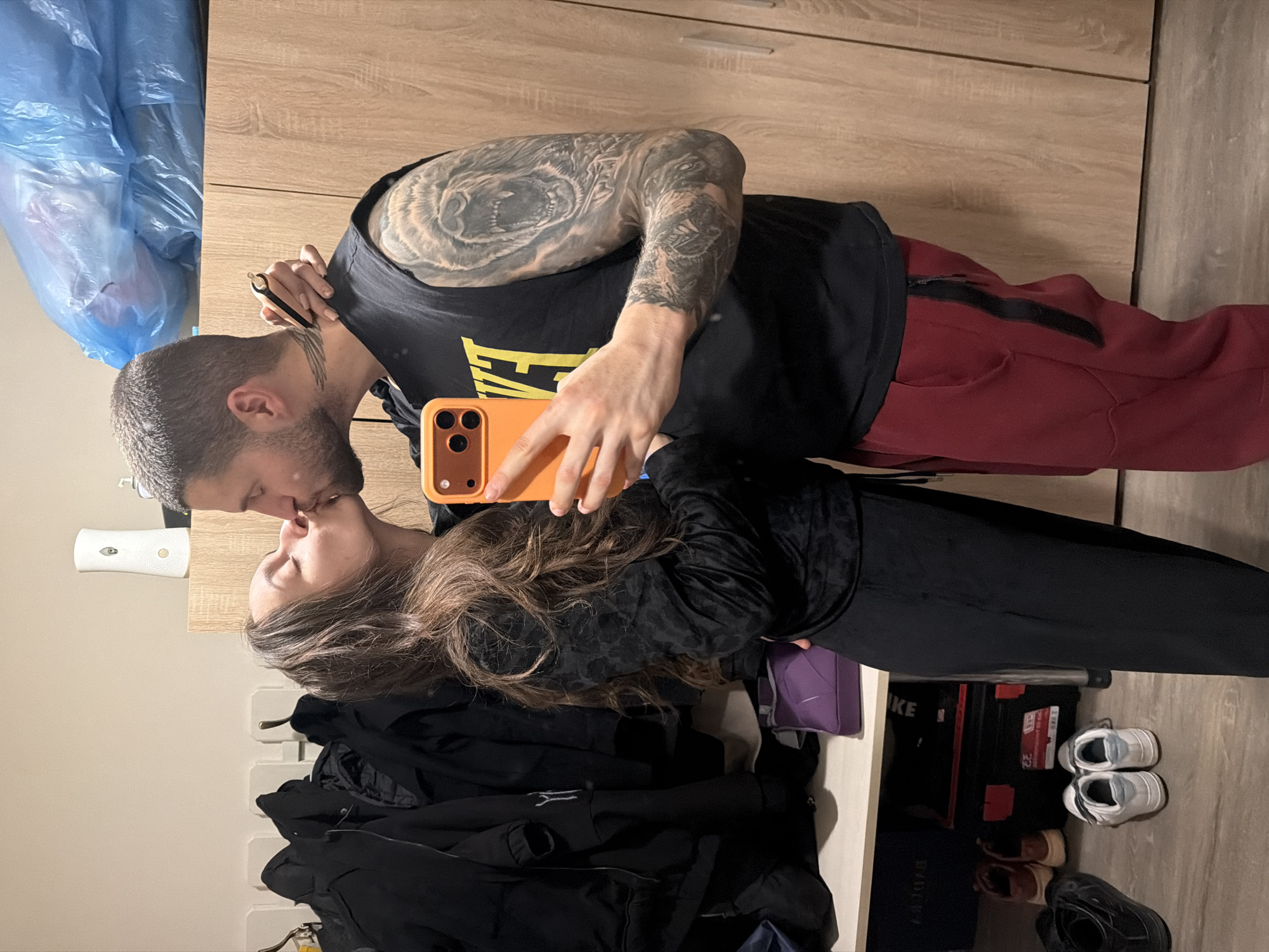 A man and woman are kissing in an indoor setting. The man has a short haircut, a beard, and extensive tattoos on his arm. He is wearing a black tank top with yellow text and red pants. The woman has long brown hair and is wearing a black outfit. The man is holding an orange smartphone, presumably taking a selfie. The background includes a wardrobe, a wall, a coat rack, and boxes of shoes. There is a blue trash bag hanging on the side. The location appears to be inside a home or apartment, but there are no specific details in the image to determine if it's in Sofia, Bulgaria, or any other location.