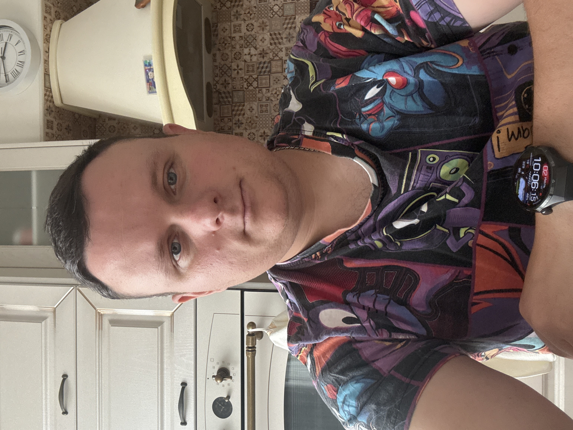 The image shows a selfie of a person in a kitchen. The person is a white male with short brown hair and blue eyes. He is wearing a t-shirt with a colorful cartoon print. He has a smartwatch on his left wrist. The background shows kitchen cabinets, a stove, and a range hood. The time on the smartwatch is 10:06. The scene is indoors, in a domestic setting. This image was taken in Kondopoga, Russia.