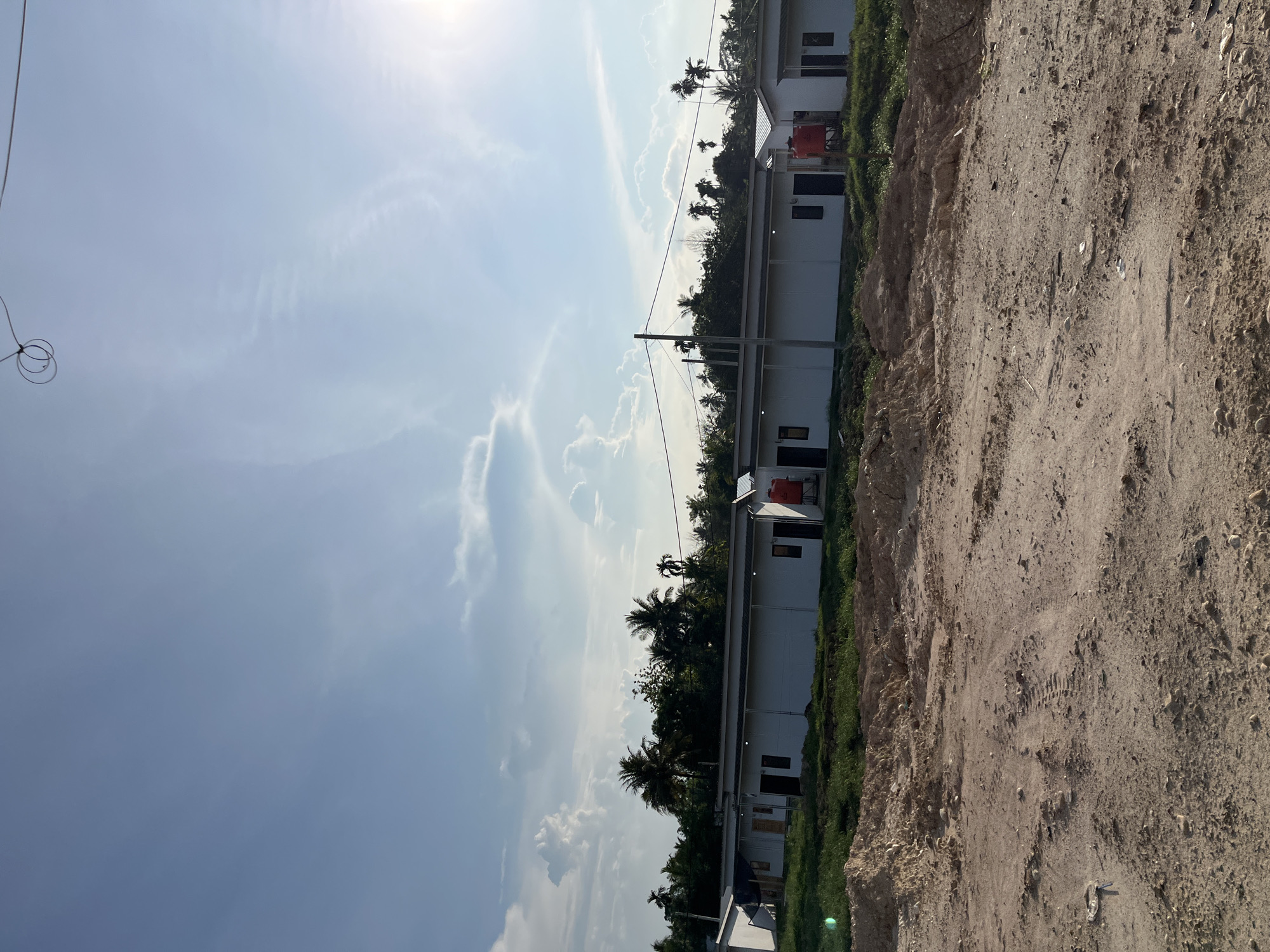 The image depicts a row of white, single-story buildings with dark-colored doors and windows, possibly residential units, under a bright blue sky with scattered clouds. In the foreground, a dirt field with tire tracks and debris suggests a construction site or undeveloped land. A few palm trees and other vegetation are visible behind the buildings. A red water tank is positioned next to one of the structures. The overall setting appears to be in a tropical or subtropical climate, consistent with the location of Alurcucur, Indonesia. There are no people or discernible activities captured in the frame.