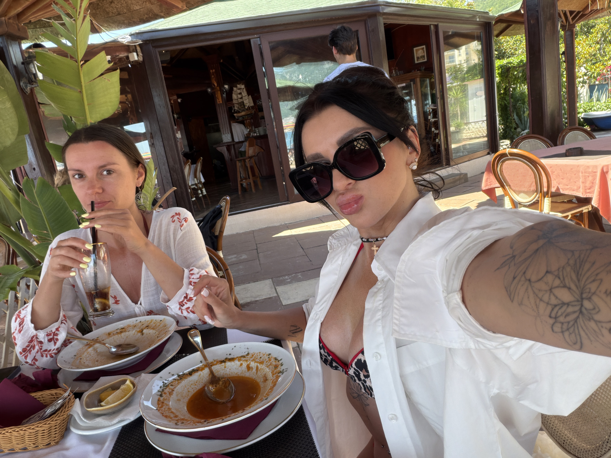 Two women are seated at an outdoor restaurant table, engaged in a selfie on a bright, sunny day in Boreti, Montenegro.
The woman on the right, taking the photograph, has dark hair styled up and wears large square dark sunglasses. She is wearing a white button-up shirt, partially unbuttoned to reveal an animal-print (leopard) bikini top and a black choker with a small black cross pendant. Visible tattoos include a large floral outline on her left arm, a smaller symbol on her right inner bicep, and a faint floral/butterfly design on her side torso. She is making a pouty expression towards the camera.
Her companion on the left has dark brown hair, pulled back, and is wearing a white V-neck top with red embroidered patterns on the sleeves. She is holding a glass containing a dark beverage with a lemon slice and a straw, looking towards the camera with a neutral expression. She has a "tree of life" earring in her visible ear.
On the dark patterned table between them are two large white bowls with gold rims, containing residual orange-colored liquid and spoons, indicating a recent meal. A small white dish with lemon wedges and a wicker basket are also present. The woman on the left has light yellow nail polish, while the woman on the right has white nail polish.
The restaurant setting features a thatched roof overhead supported by wooden beams, and large green tropical plant foliage partially separates the two women. Behind them is a glass-fronted building, part of the restaurant, with interior wooden decor, chandeliers, and a model ship visible. A man is partially visible inside the building. Reflections in the glass show a body of water and distant mountains, consistent with a coastal environment. To the right, additional outdoor tables with pink tablecloths and wooden chairs are arranged. The ground is paved with stone or tile.