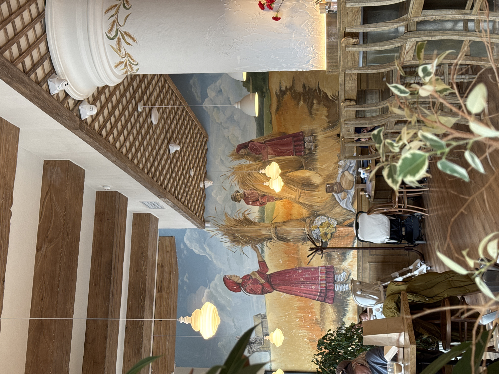 The image depicts the interior of a restaurant in Izhevsk, Russia. The decor features folk-art elements, including a mural of peasants harvesting grain, wooden beams, and decorative trellis work. Tables and chairs are visible, some occupied by patrons. A baby stroller is present. Foliage from plants obscures some parts of the scene.