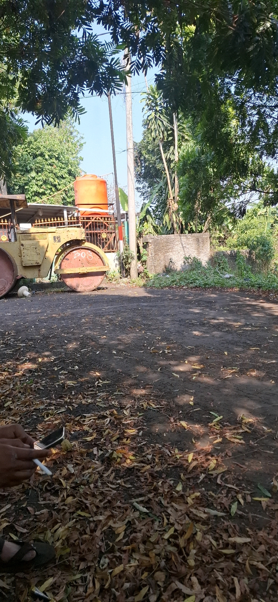 A vintage yellow steamroller sits on a dirt road in a residential area of Cilaku, Indonesia. The steamroller, showing signs of rust and wear, is positioned near a bright orange water tank and a dilapidated fence. Lush green trees and foliage surround the scene, with fallen leaves scattered across the ground. In the foreground, a person's hand is visible, holding a lit cigarette and a mobile phone, suggesting a moment of pause. The image captures a quiet, sun-drenched afternoon, with shadows cast by the trees dappling the ground.