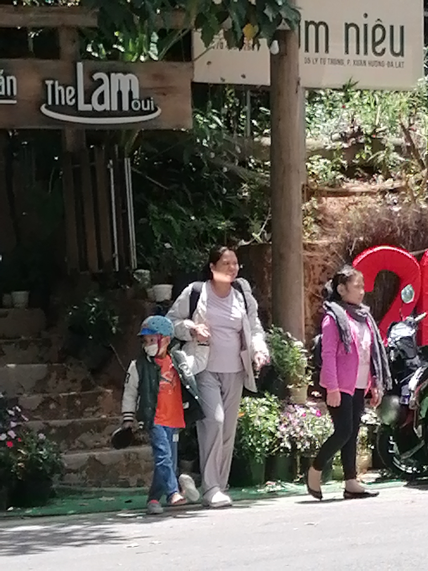 The image shows three people walking on a paved surface in front of a building or establishment with signage. On the left, a young boy wearing a blue hat, a mask, a green and white jacket over an orange shirt, and blue jeans is walking away from the camera. He is holding a dark-colored object. To his right, a woman in a light-colored jacket and pants is walking past. To the right of the woman, a young girl with her hair in a ponytail, wearing a pink jacket and black leggings, is walking away from the camera. She has a dark scarf around her neck and is carrying a backpack. Behind the people, there are steps leading up to a wooden structure with signage. One sign reads "an The Lam oui". Another sign further back has Vietnamese text, with "m niêu" prominently visible, and smaller text below that includes "05 LY TU TRONG, P. XUAN HUONG, DA LAT". This indicates the location is 05 Ly Tu Trong Street, Xuan Huong Ward, Da Lat City, Vietnam. There are plants and foliage in the background, and a large red inflatable object is partially visible on the far right. The lighting suggests it is daytime, with shadows cast on the pavement.