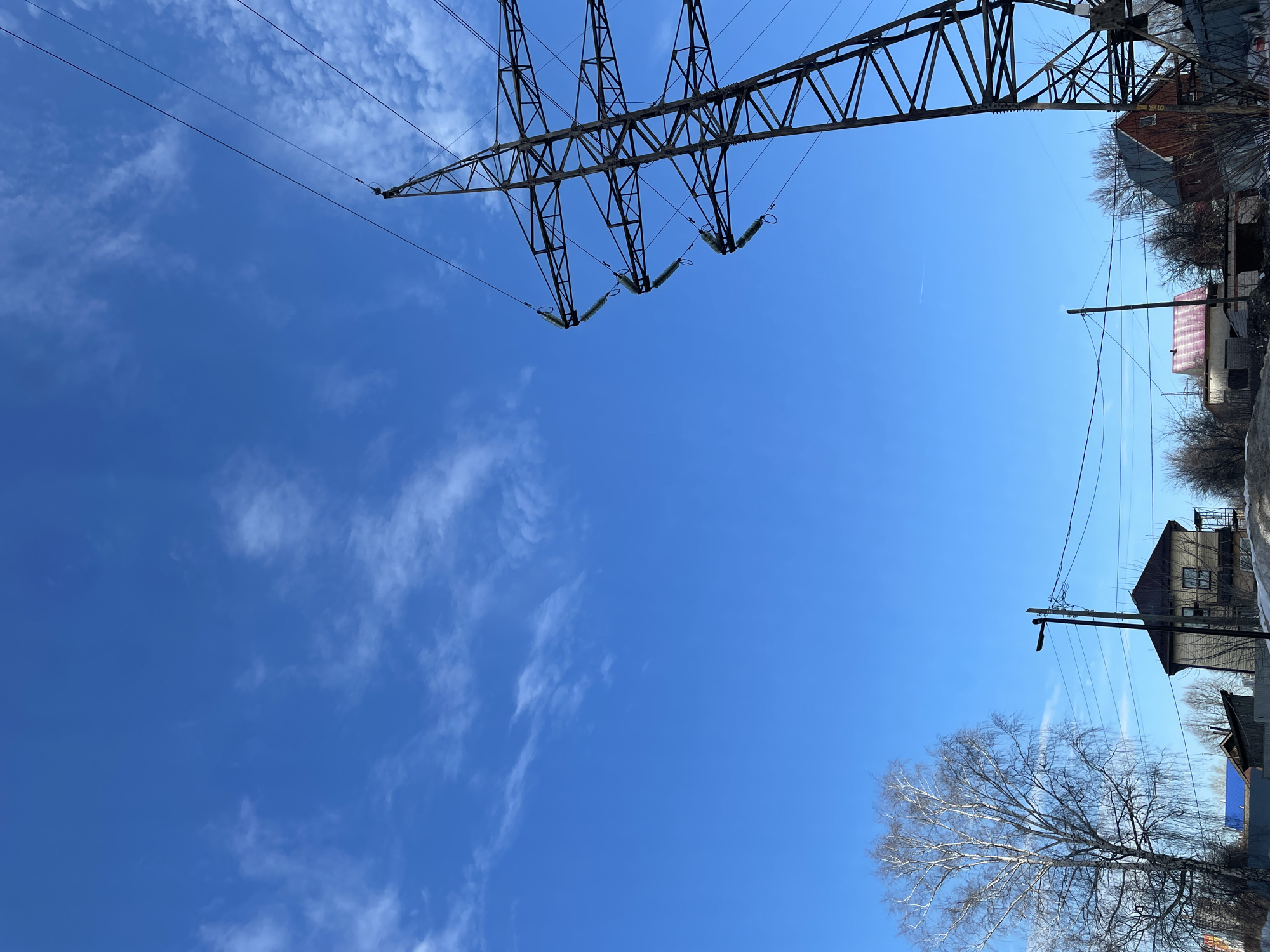 The image depicts a daytime scene in a semi-urban residential area of Izhevsk, Russia. The sky is bright blue with scattered, wispy cirrus clouds. A large metal lattice power transmission pylon occupies the right side of the frame, extending from the lower right to the upper right quadrant. Multiple overhead power lines are suspended from the pylon via sequences of disc insulators. In the left foreground, a bare deciduous tree, characterized by light-colored bark and intricate branch patterns, stands prominent. Below the horizon, several residential buildings are visible, including a two-story light-colored house with a dark roof. A wooden utility pole, equipped with a streetlamp and multiple electrical wires, is positioned in front of this house. Further right, partially obscured by the pylon, additional smaller houses with varying roof colors (e.g., red, blue) are observed, with construction scaffolding adjacent to one structure. The ground consists of bare earth with visible patches of snow. No individuals are present, and no actions or interactions are depicted.