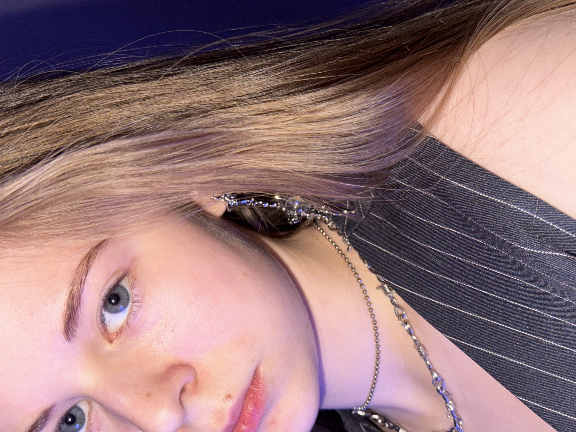A close-up portrait shows the upper body of a young person, likely female, with fair skin and light brown hair that has lighter blonde highlights. The subject is positioned diagonally in the frame, looking directly at the viewer with piercing blue-grey eyes and a neutral, contemplative expression. Her long hair, with visible darker roots, flows towards the upper left, set against a solid dark blue background.
She is wearing a dark blue or black pinstripe garment, possibly a jacket or shirt, visible on the right side of the image. Decorative silver-toned jewelry adorns her: a multi-chain earring or ear cuff on her visible left ear, and another finer silver-colored chain draped around her neck and chest area.
The lighting on her skin has a distinct pinkish and purplish hue, suggesting artificial light sources. While no specific location cues are visible within the image itself, the context provided indicates the photograph was taken in Perm, Russia. No visible text is present in the media file.