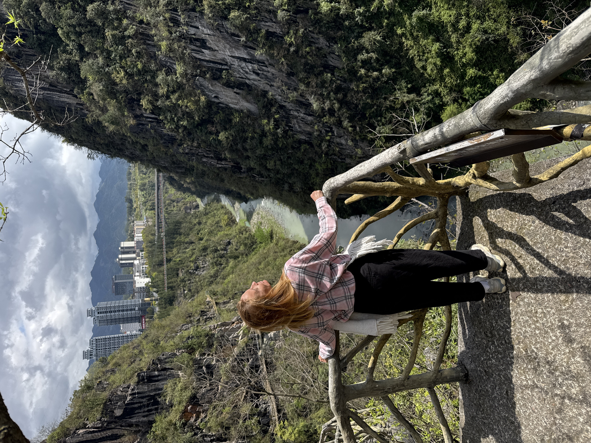 A young woman stands on a concrete platform overlooking a river and a cityscape, in Qianjiang, China. She is wearing a pink and white plaid shirt and black pants, with white sneakers. She leans against a rustic railing constructed to look like tree branches. The platform is bordered by a sheer rock cliff covered in green foliage on one side, and on the other side is a view of a river winding between hills, with tall buildings in the distance. The sky is cloudy.