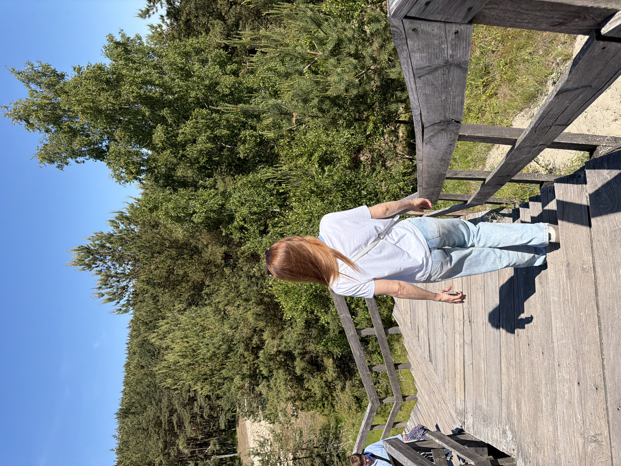 In Morskoye, Russia, a woman with auburn hair walks down a wooden staircase, her back to the camera. She wears a white t-shirt and light blue jeans. To her left and slightly ahead, another person in a blue and white checkered shirt descends the stairs. The staircase is set against a backdrop of dense green coniferous and deciduous trees under a clear blue sky. The wooden structure shows signs of weathering.