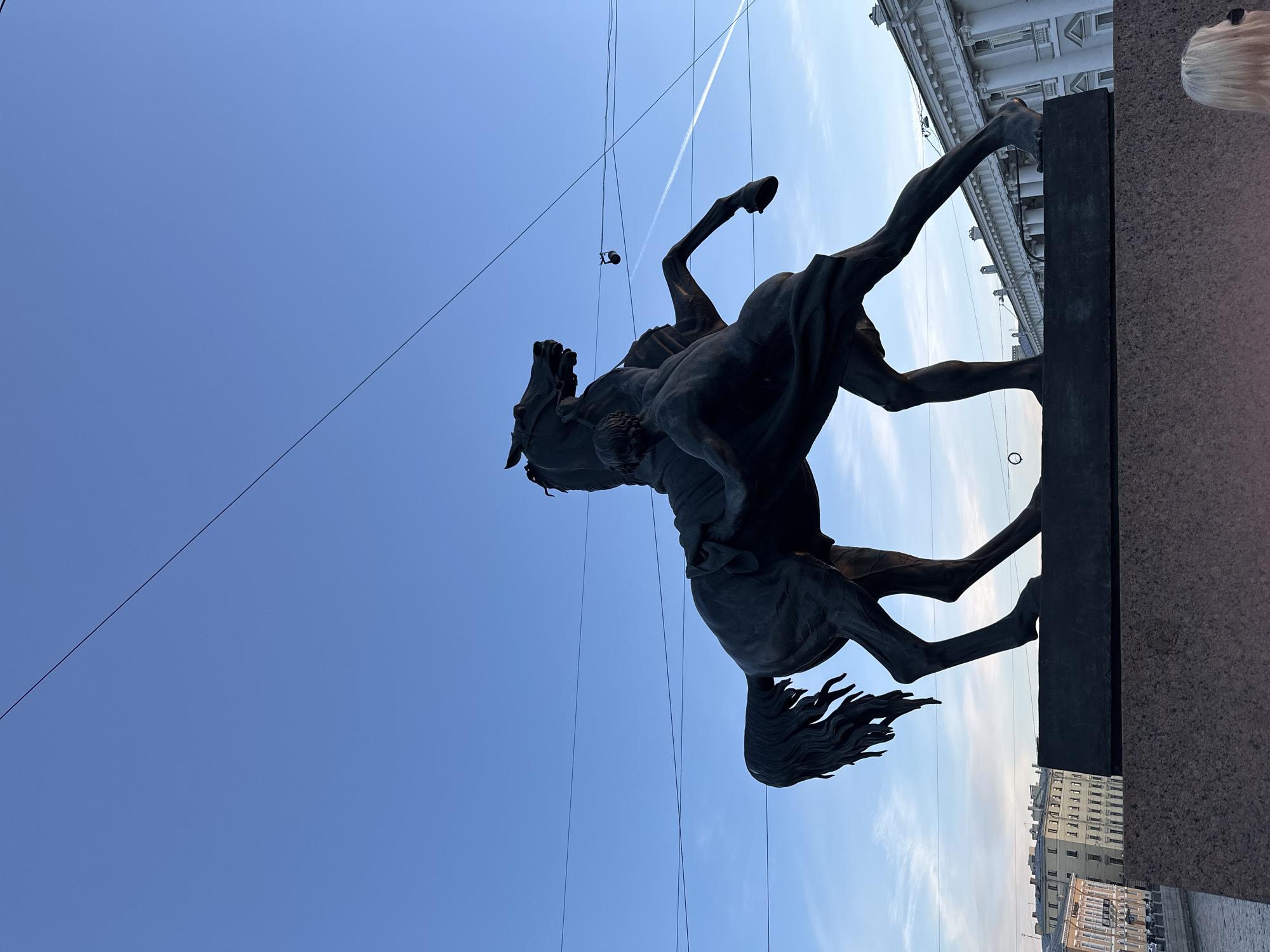 The image captures the Bronze Horseman statue in Saint Petersburg, Russia, viewed from a low angle. The statue, a bronze equestrian monument of Peter the Great, is the dominant subject, rendered in silhouette against a blue sky with faint contrails. Utility lines stretch across the sky. The statue is positioned on a dark plinth atop a granite base, with a portion of a building visible in the background, suggesting a city setting. The overall impression is a dramatic and upward-looking perspective on this iconic monument.