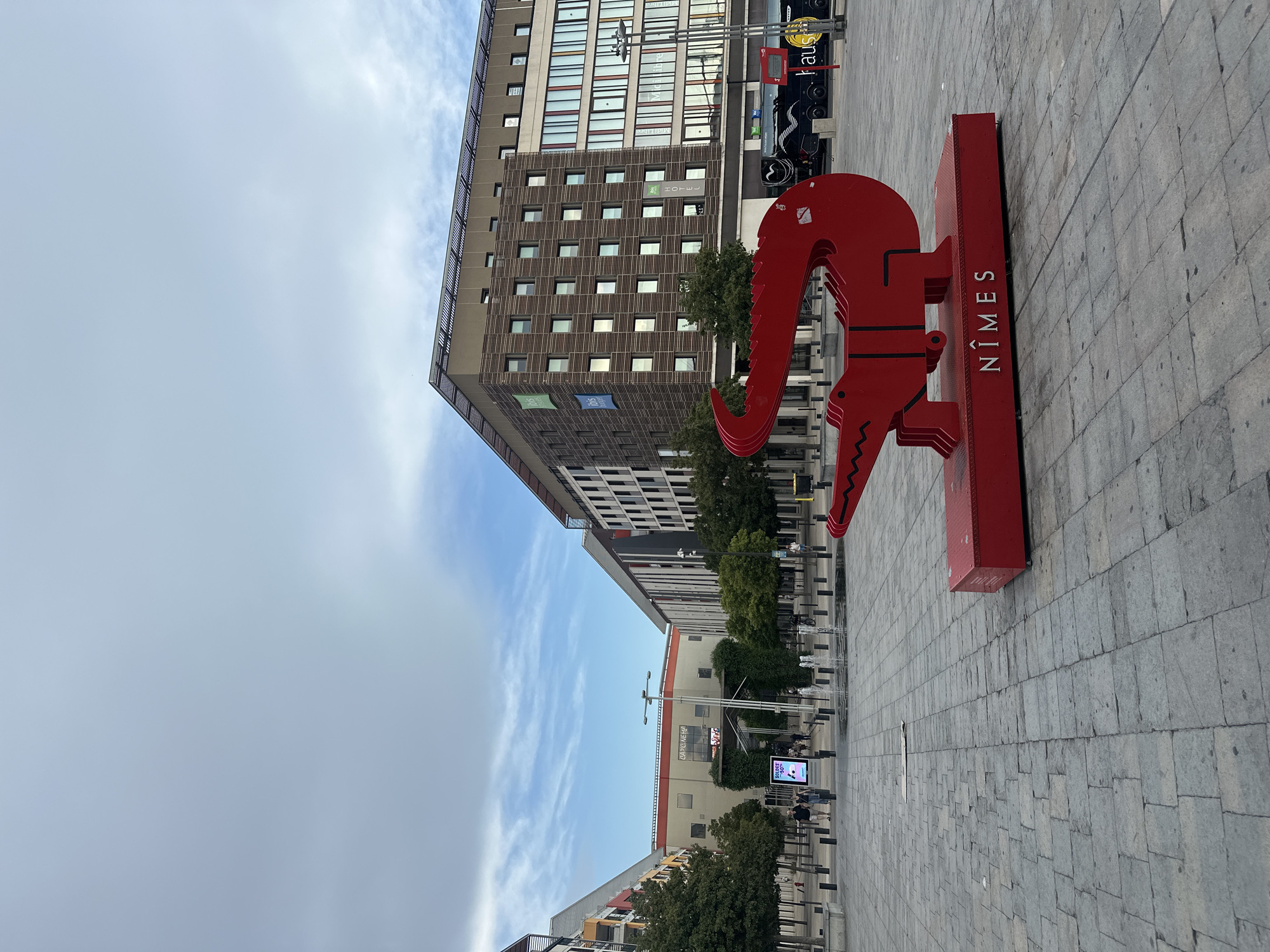A bright red, stylized sculpture of a crocodile on a red platform is positioned in a paved plaza in the city of Nîmes, France. The sculpture has the word "NÎMES" spelled out in white block letters on the front of its platform. Behind the sculpture, modern buildings with many windows line the plaza, including a brown building with solar panels on its roof and several green "ibis Hotel" logos visible. To the left of the sculpture, more buildings are visible, as well as trees and a fountain. Several people are walking in the plaza, and a black bus with white lettering is parked in the background. The sky above is partly cloudy.