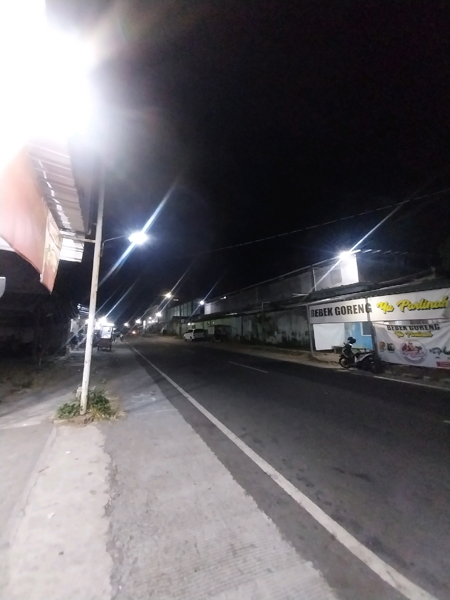 The image shows a street scene at night in Payaman, Indonesia. A bright street light illuminates the foreground on the left. A sidewalk runs along the left side of the street, with some vegetation growing near a utility pole. A food cart is parked on the sidewalk. Across the street, there is a building with signs that read "BEBEK GORENG Yu Partinah" and "BEBEK GORENG Yu Partinah" on a banner. A motorcycle is parked in front of the building. The street itself is paved and has a white line marking the lane. The sky is dark, indicating it is nighttime.