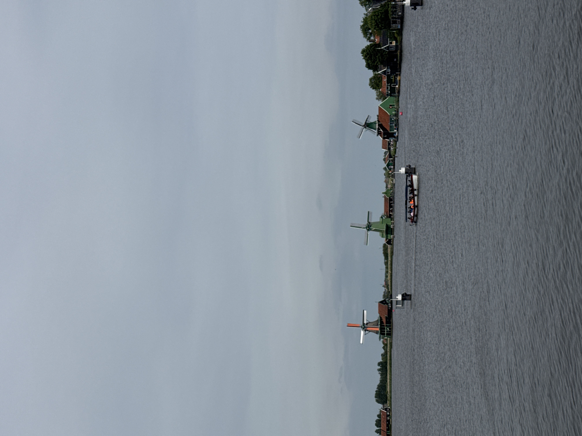 The image captures a view of Zaanse Schans, Zaandijk, Netherlands. Several traditional Dutch windmills stand along a waterfront. A boat carrying passengers floats near the windmills. The sky is overcast.