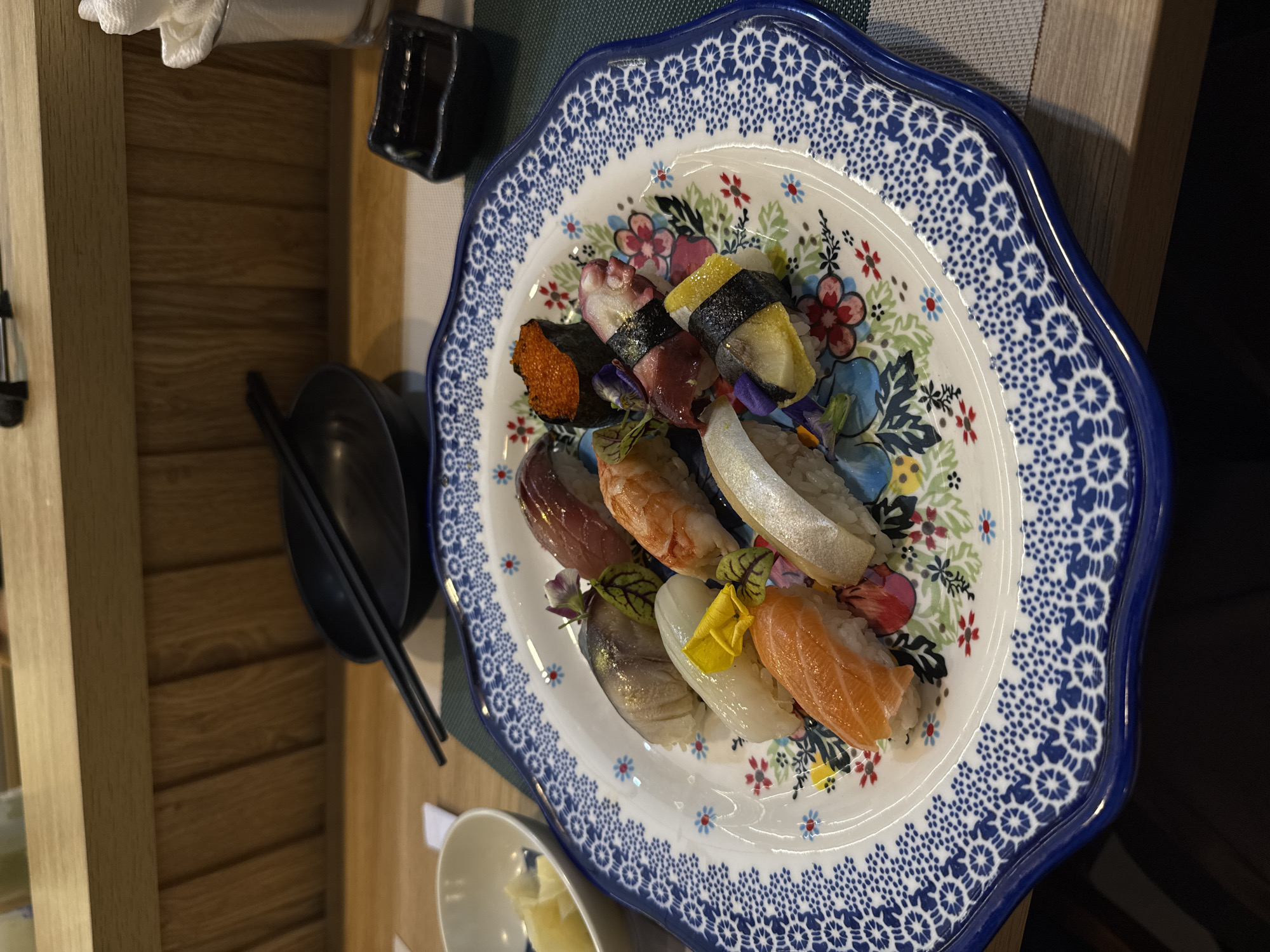 A plate of sushi sits on a table in a restaurant, possibly located in Nha Trang Ward, Vietnam. The plate is decorated with blue and white patterns and floral designs. Assorted sushi pieces, including nigiri and possibly some maki, are artfully arranged on the plate. Some pieces feature salmon, tuna, shrimp, and possibly other types of seafood. A pair of chopsticks rests beside the plate, and other small dishes and table accessories are visible. The table appears to be wooden. The lighting is soft, suggesting an indoor setting.