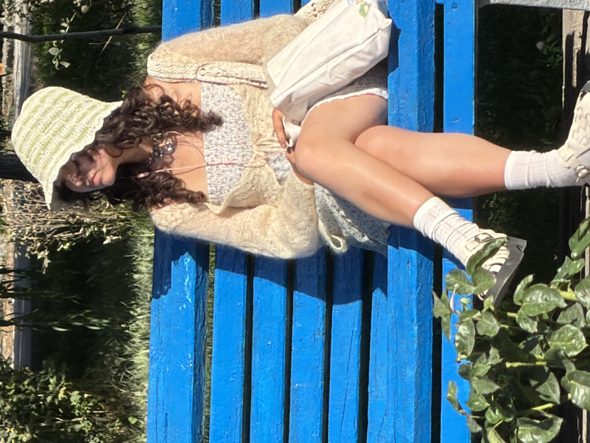 A person is sitting on a bright blue wooden park bench, facing right, with their legs crossed. They are wearing a light-colored crocheted bucket hat, a floral-patterned top, a light tan cardigan, a light-colored skirt, white socks, and sandals. A white tote bag is resting on the bench next to them. The background shows greenery, including a rose bush in the foreground, and the setting appears to be a park or outdoor area in daylight.