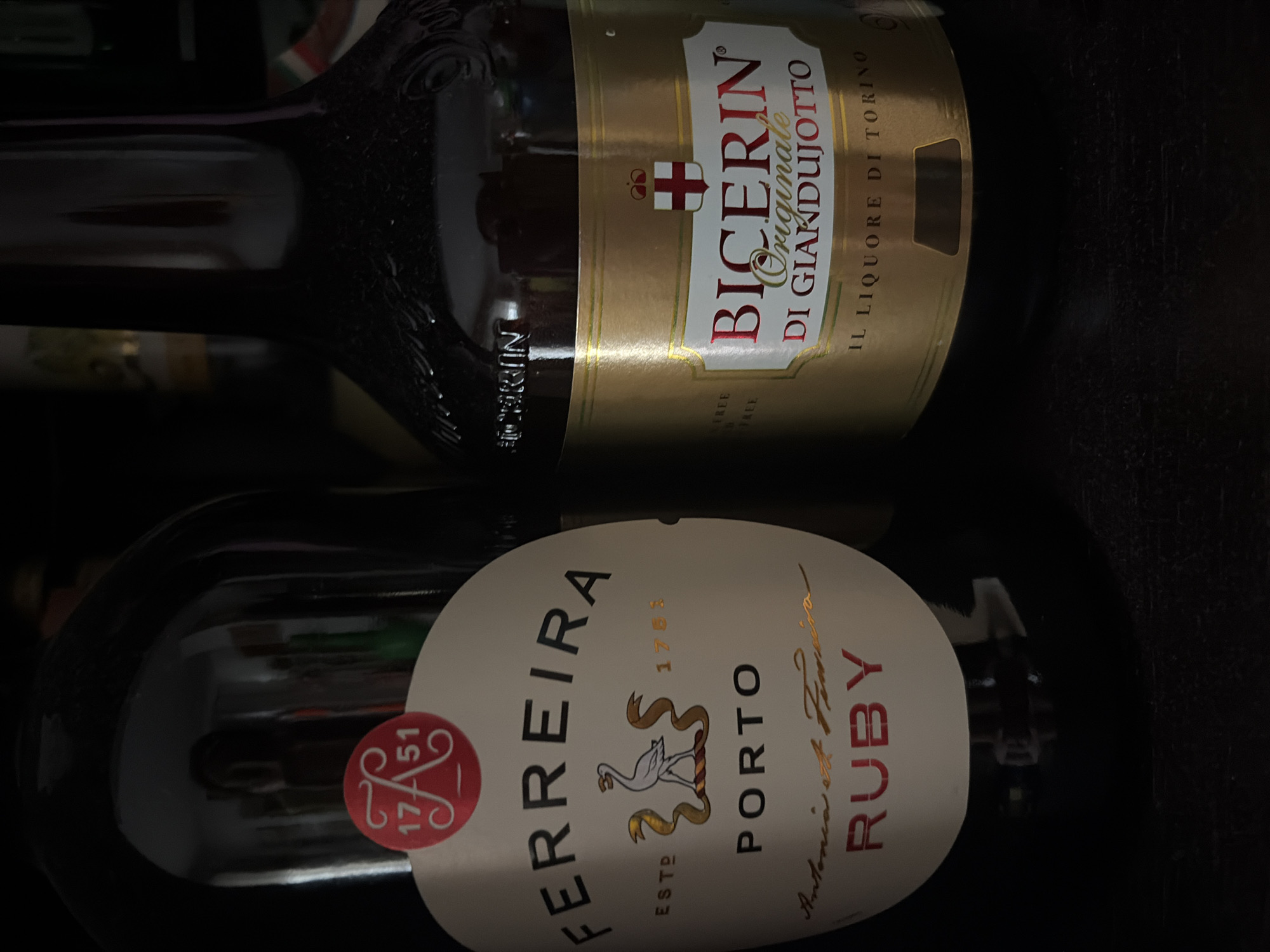 The image shows two bottles of alcohol in a dark environment, with a faint reflection of a person in the background. The bottle on the left has a white label with "FERREIRA PORTO" written in black. Below that, in a cursive font, it says "Antonia et Ferreira" and "RUBY" in red. There is also a red circular emblem with "1751" inside. The bottle on the right has a gold label with "BICERIN ORIGINALE DI GIANDUJOTTO" written in red and green. Below that, it says "IL LIQUORE DI TORINO". The image does not provide any information about the city of Bari, Italy.