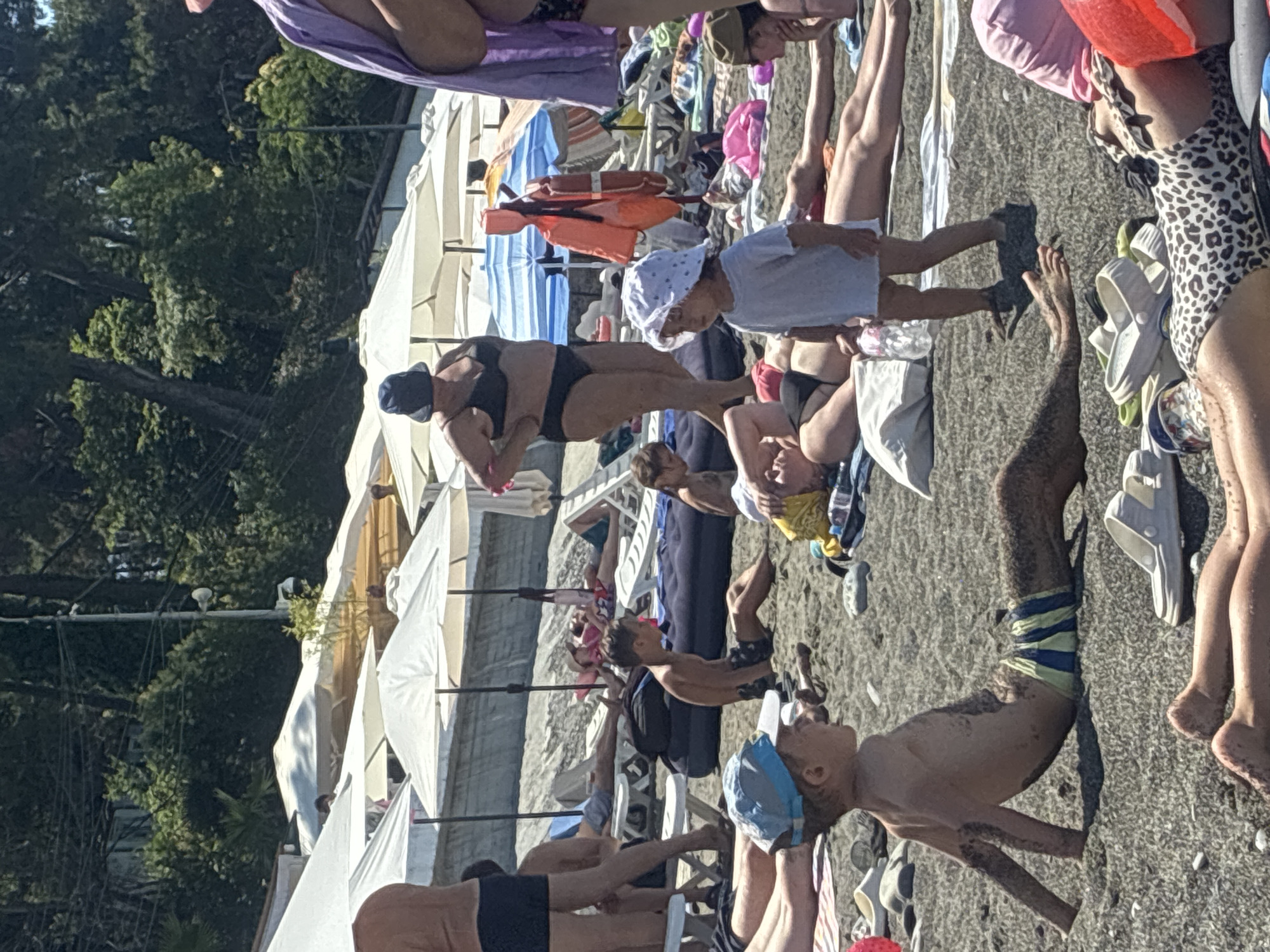 This is a photograph taken on a sunny day at a crowded beach in Sochi, Russia. The beach is covered in dark grey pebbles and sand. In the foreground, a young boy wearing a blue and yellow striped swim trunk and a blue cap sits on the ground, covered in sand. To his right, another child wearing a white hat and shirt stands. Several adults are sunbathing on lounge chairs or lying on towels in the background, some partially obscured by beach umbrellas. The umbrellas are predominantly white and yellow, providing shade for beachgoers. A woman in a black bikini and a dark blue sun hat stands in the mid-ground. Orange life vests are visible hanging from a structure. The sea is not visible in the frame, but the presence of umbrellas and people suggests it is nearby. The overall scene depicts a typical summer day at a popular resort beach.
