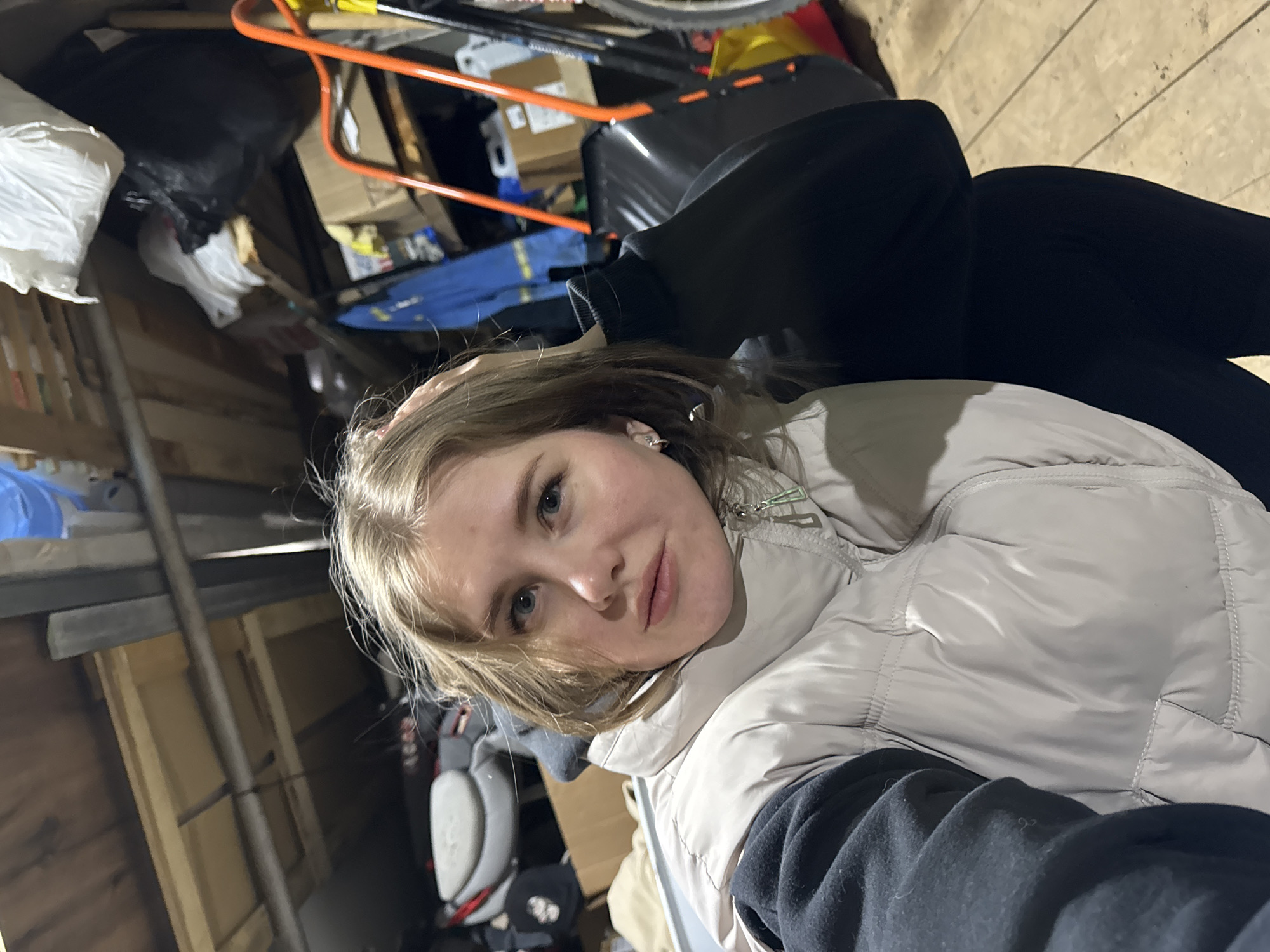 A woman wearing a light-colored puffer jacket and black pants is sitting or lying in what appears to be a cluttered garage or storage area. The background contains various items, including cardboard boxes, a black trash bag, tools with orange handles, and other miscellaneous objects. The walls are made of wood, possibly unfinished planks. The location is unspecified, but the user has declared it is in the city of Gornyi (Dvurechenskoe S/P), Russia.