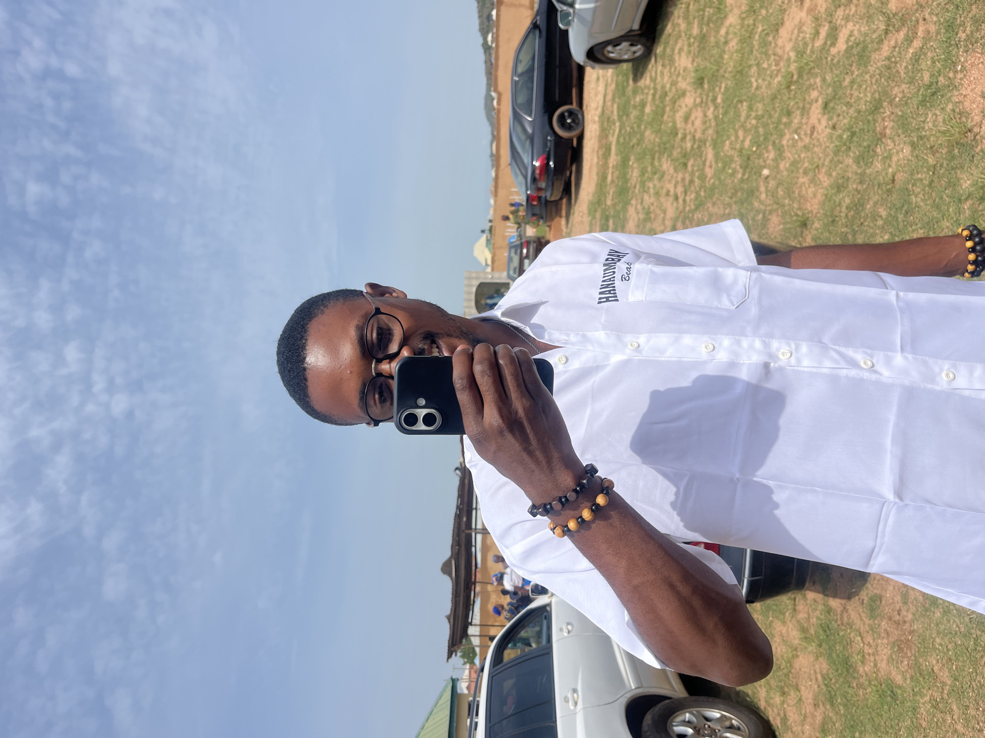 A man with dark skin, glasses, and short black hair is taking a selfie outdoors. He is wearing a white short-sleeved shirt with a pocket on the left side that has the text "HANAUMBAY Beat" embroidered in dark blue. He is holding a black smartphone in his right hand and is wearing two beaded bracelets on his left wrist. The bracelets consist of dark and light brown beads. The background shows a grassy area with several cars parked, including a white SUV on the left, two black sedans in the middle, and a silver sedan on the right. In the distance, there are some buildings and structures, and the sky is blue with scattered white clouds. The lighting suggests it is daytime. The location is identified as the city of Jos, Nigeria.