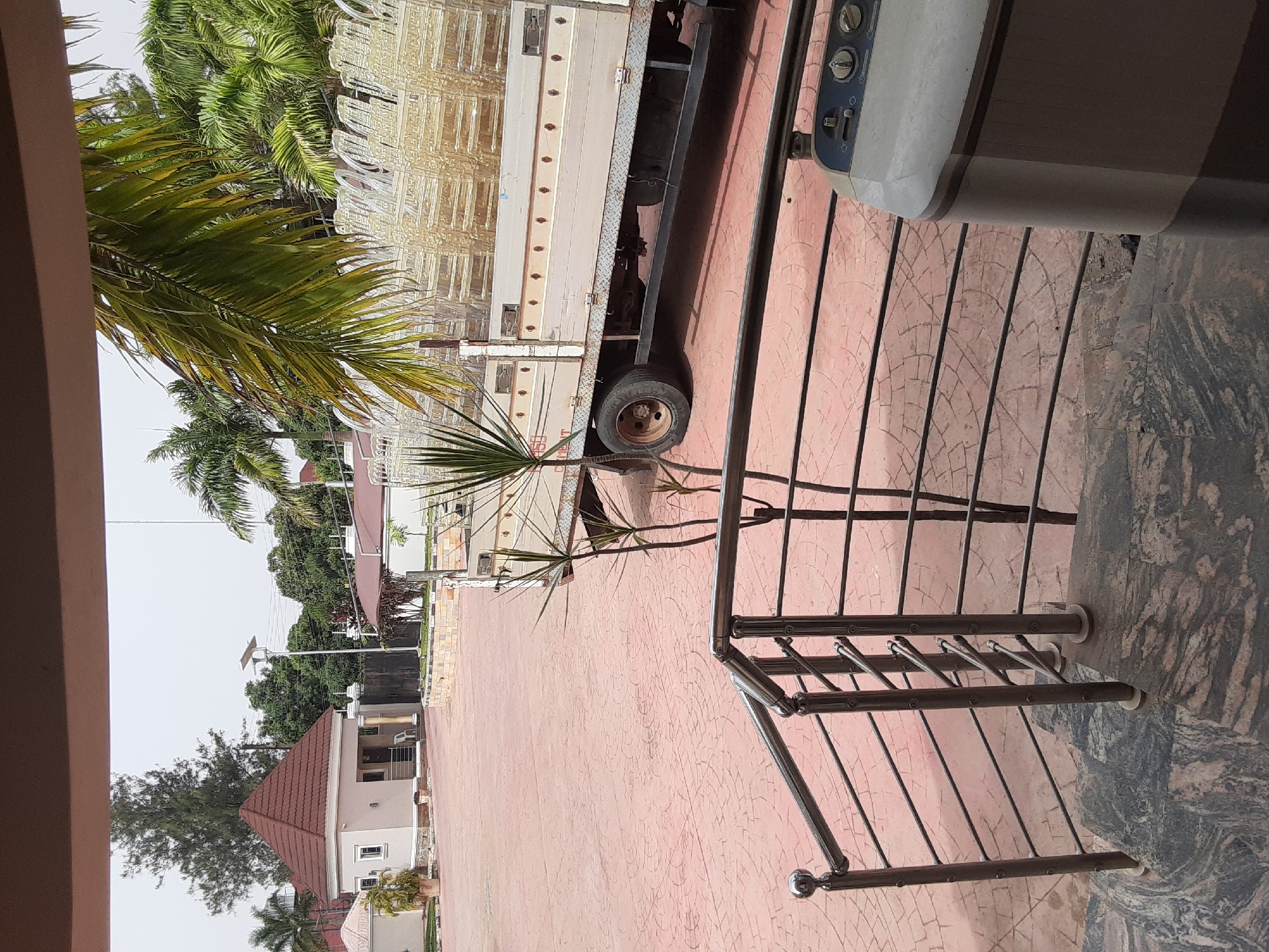 The image shows an outdoor scene from a location within Gura Top, Nigeria. The foreground features a stainless steel railing on a patio with red paved bricks and a marble-like stone section. Adjacent to the railing is a possibly malfunctioning appliance. A flatbed truck is parked on the paved area, loaded with many white chairs. A palm tree is on the left. Buildings and trees are visible in the background. The sky is overcast.