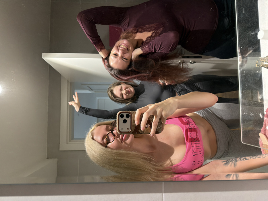 An image captured in an interior room, likely a residential bathroom, in Belgrade, Serbia, depicts three adult women posing for a mirror selfie. The blonde woman in the foreground wears spectacles, a pink crop top, and grey leggings, holding a smartphone with a textured case to capture the reflection. A tattoo is visible on her left forearm. To her right and slightly behind, a second woman, dressed in a black long-sleeved top and dark trousers, smiles with narrowed eyes and her right arm extended upwards. On the far right, a third woman, wearing a maroon long-sleeved top with lace trim, is observed smiling widely, tilting her head, and grasping her hair with both hands. All three subjects display expressions of amusement. The background, reflected in the mirror, shows light grey walls and a white doorway. The bottom edge of the mirror reveals a counter surface with a folded reddish towel, a small decorative object, and a white cylindrical container.