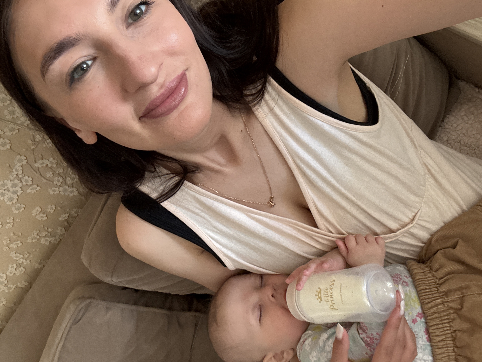 A woman is holding a baby in her arms and feeding it a bottle. The baby is lying on its back, with the bottle held up to its mouth. The bottle has the words "Little Princess" on it. The woman is looking at the camera and smiling. The background is a couch or bed. The image was not taken in Sochi, Russia.