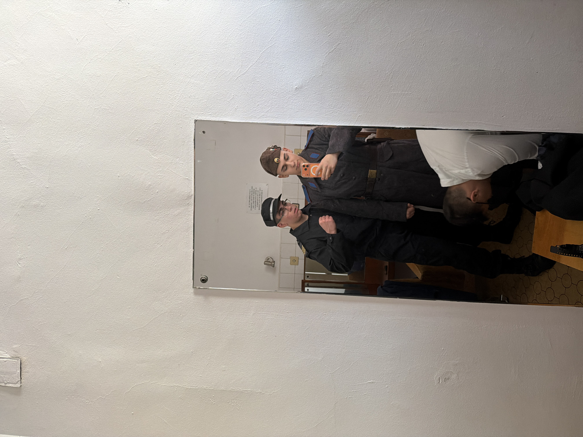 A mirror hanging on a white wall reflects three people. Two of the individuals, dressed in what appears to be military or law enforcement uniforms, are posing for a selfie; one holds a cell phone. The third person is partially visible, with only their head and shoulders appearing in the mirror's reflection as they stand next to the other two. The backdrop reflected in the mirror suggests an interior space, potentially a bathroom or locker room, with light-colored walls and visible fixtures. The location is not discernible from this image.
