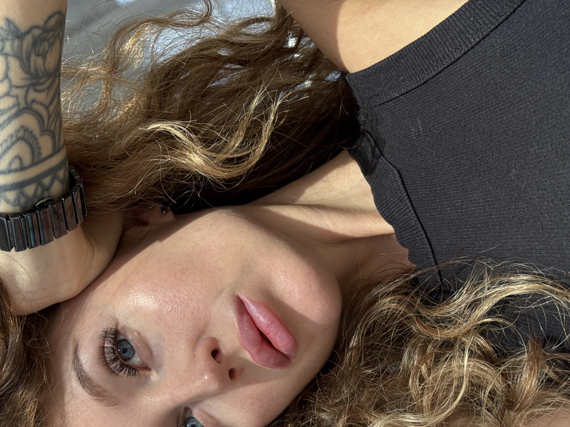 This close-up portrait, taken in Chaikovsky, Russia, features a woman lying down, illuminated by bright natural light, suggesting daytime. The primary subject's head and upper body are angled, with her gaze directed towards the viewer.
She has fair skin, blue eyes, and long, dark eyelashes, which appear to be extensions. Her lips are full with a rosy hue, possibly from lip blush or filler. Her light brown to blonde curly hair is spread around her. A large, dark gray tattoo with floral or paisley patterns is visible on her left inner forearm, near a dark, segmented metal watch on her wrist. She is wearing a simple black top with a ribbed texture, on which some light fibers are visible.
The lighting creates soft shadows and highlights on her face and hair. The background is bright and out of focus, providing no specific environmental details. There is no visible text in the image. The overall mood is calm and contemplative.