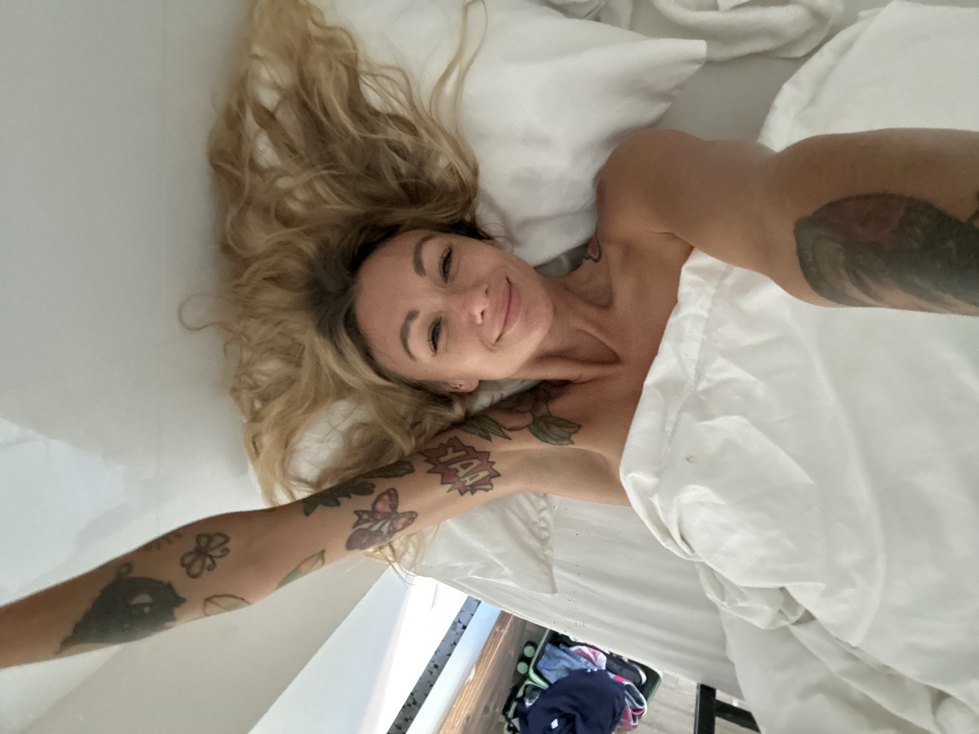 A woman with blonde hair and tattoos on her arm lies in bed, smiling at the camera. Her right arm is raised above her head. She is covered by a white duvet. In the background, a suitcase with clothes is visible.