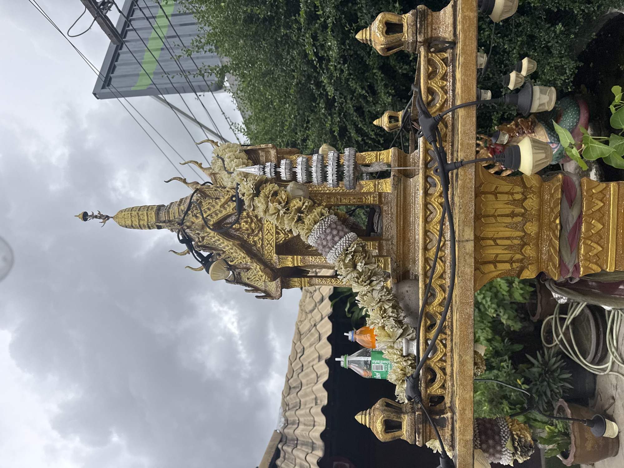 The image shows a golden spirit house, likely a traditional Thai San Phra Phum, situated outdoors. The spirit house features intricate golden detailing, including tiered roofs, decorative finials, and ornate pillars. It is adorned with garlands of light-colored flowers and a silver tiered ornament hanging from the side. Two bottles, one green and one orange, are placed on the structure behind the garlands. The spirit house is illuminated by a string of small, decorative lights. In the background, a tiled roof of a building is visible to the left, and lush green trees and bushes frame the right side of the scene. Overhead, a cloudy, overcast sky suggests a humid or rainy day. A portion of a grey, industrial-looking structure with green stripes and electrical wires is also present in the upper right corner. The ground beneath the spirit house is not clearly visible, but potted plants and a coiled green hose suggest an outdoor, possibly residential or commercial, setting. No people or animals are depicted in the image. The presence of the spirit house indicates a religious or spiritual element within the urban landscape of Chalong, Thailand.
