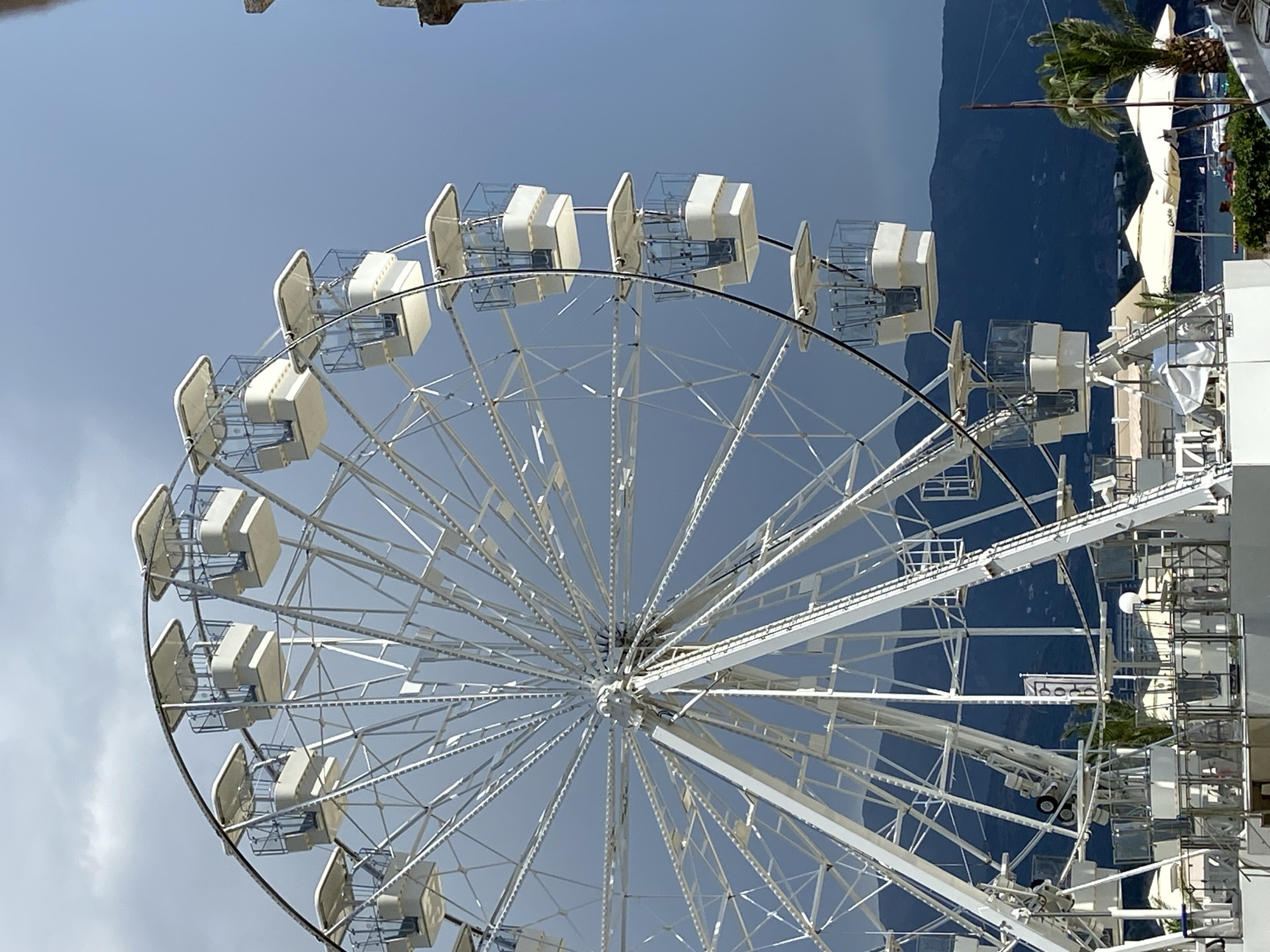 The media file shows a large white Ferris wheel with several gondolas. The Ferris wheel is the primary subject, taking up most of the frame. The gondolas are enclosed and appear to be designed for passengers. The Ferris wheel is set against a backdrop of a clear blue sky with some light clouds. In the distance, a dark, mountainous landscape is visible, suggesting a coastal or hilly region. There are also some structures visible behind and to the right of the Ferris wheel, including white umbrellas, some greenery, and buildings that appear to be part of a resort or entertainment area. The lighting suggests it is daytime, likely a sunny day. No people are clearly visible, though the presence of the Ferris wheel and resort-like structures implies a recreational setting. There is no visible text in the media file. The orientation of the image is rotated. The location context of Budva, Montenegro, aligns with the visual cues of a coastal town with mountainous terrain and a clear sky, often associated with tourist attractions like Ferris wheels.