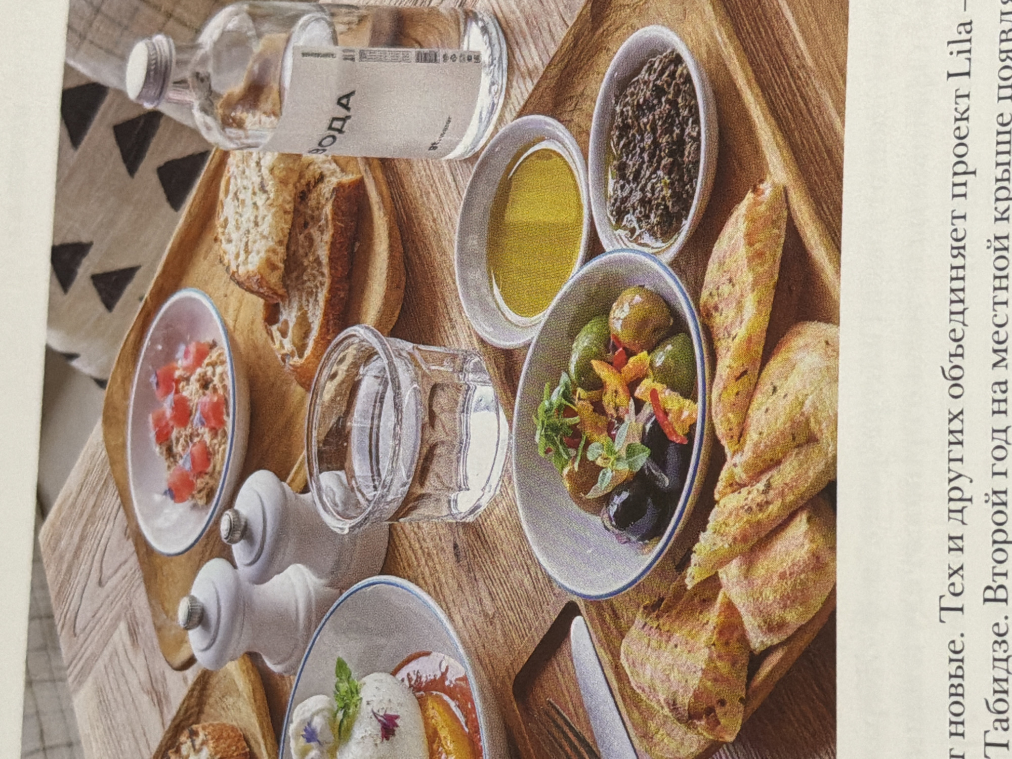 Here's a detailed description of the image:
**General Description:**
The image captures a selection of food and drinks arranged on a wooden serving board or tray. The setup suggests a meal or snack intended to be shared.
**Objects and Food Items:**
1. **Water Bottle:** A clear glass bottle, labeled "ВОДА" (Water in Russian).
2. **Bread:** Sliced brown bread, possibly whole wheat, is placed near the water bottle.
3. **Bowls:**
* A bowl with diced tomatoes in a dressing.
* A bowl with a cream-colored food item, garnished with herbs and edible flowers.
* A bowl filled with olives and other preserved vegetables, decorated with herbs.
* A bowl with dark olive tapenade.
4. **Olive Oil:** A small bowl filled with olive oil.
5. **Glass:** A clear glass.
6. **Salt and Pepper Shakers:** White salt and pepper shakers.
7. **Bread Pieces:** Several pieces of light-colored, possibly artisanal bread are near the olive bowl.
8. **Knife and Fork:** The utensils on the right indicate that it is a table setting.
**Environment:**
The scene is set on a wooden surface, likely a table or countertop. Part of a newspaper or magazine is visible, with text in the Russian language.
**Possible Location:**
Based on the text in Russian, the location is likely Moscow, Russia. The text mentions "Табидзе," which is a Georgian surname, and refers to "новый проект Lila," suggesting a new project or restaurant, or initiative.