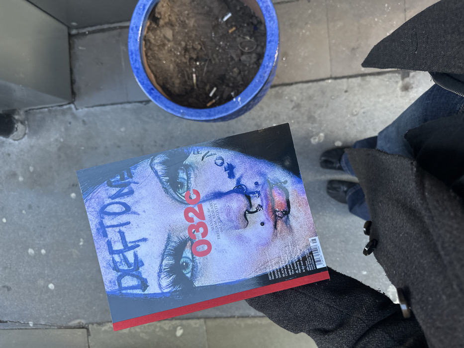 An overhead photograph depicts a person standing on grey paving slabs in the city of London, United Kingdom. The individual's lower torso, clad in blue jeans, and black split-toe shoes are visible, along with a dark coat sleeve on the left. The person holds a copy of "032c" magazine. The magazine's cover displays a stylized, purplish-blue portrait of a face featuring prominent eyelashes, dark eye makeup, and multiple facial piercings, with "DEFTONE" scrawled across the forehead and "032c MANUAL for FREEDOM, RESEARCH and CREATIVITY" printed below. To the right of the magazine, a blue ceramic pot contains dark soil and several scattered white cigarette butts, indicating its use as an ashtray.