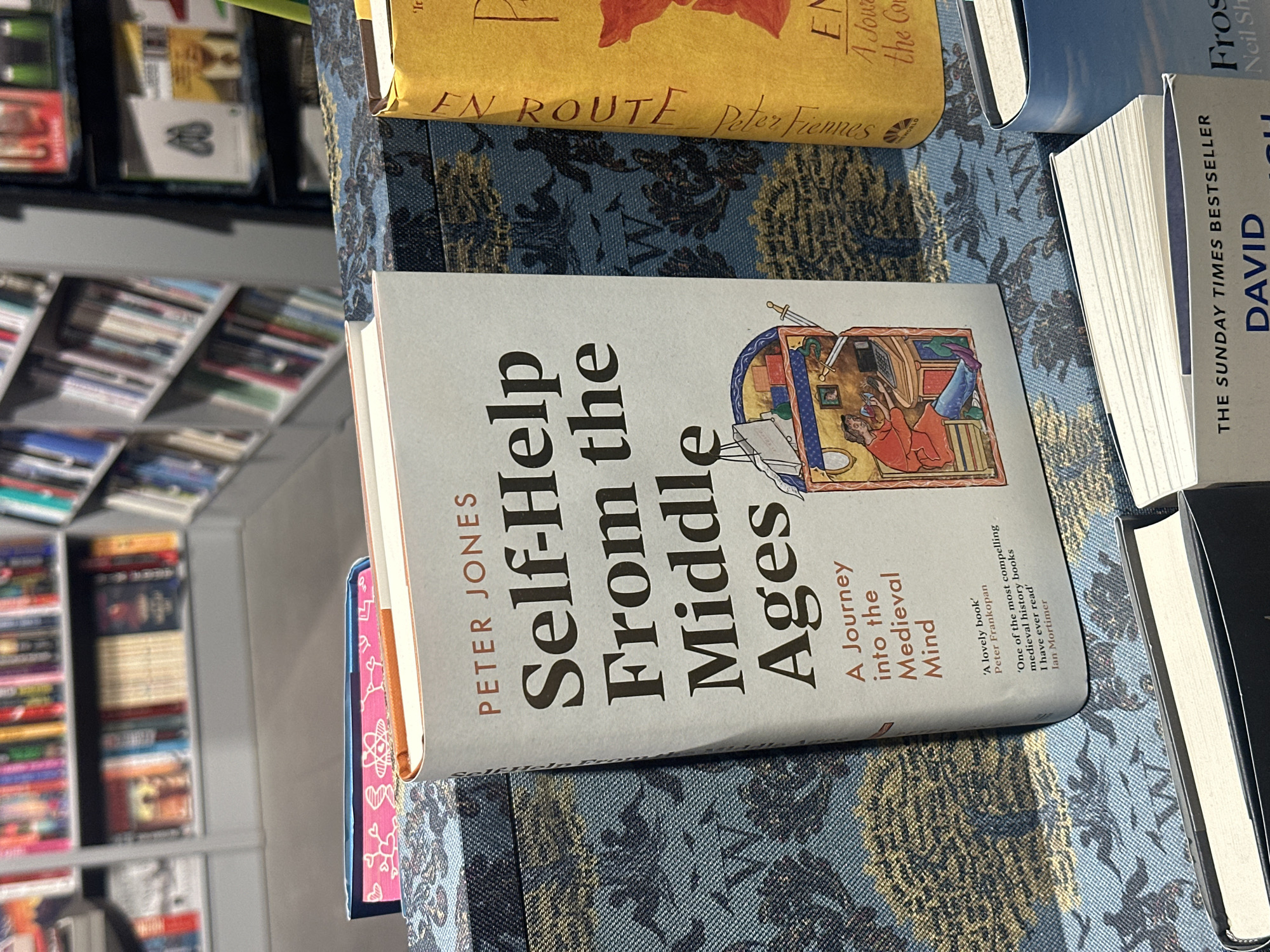 The image shows a close-up of several books placed on a surface with a patterned, blue fabric. The main book in focus is titled "Self-Help From the Middle Ages" by Peter Jones. It features an illustration on the cover depicting a medieval scene. Adjacent to it are other books; one with a yellow cover with the title "EN ROUTE Peter Fiennes" and another with the title "DAVID", identified as "THE SUNDAY TIMES BESTSELLER". In the background to the left, a bookshelf is visible, densely packed with various books. The image provides no indication of a specific city or location, so it is impossible to place the scene in "Unknown, Unknown".