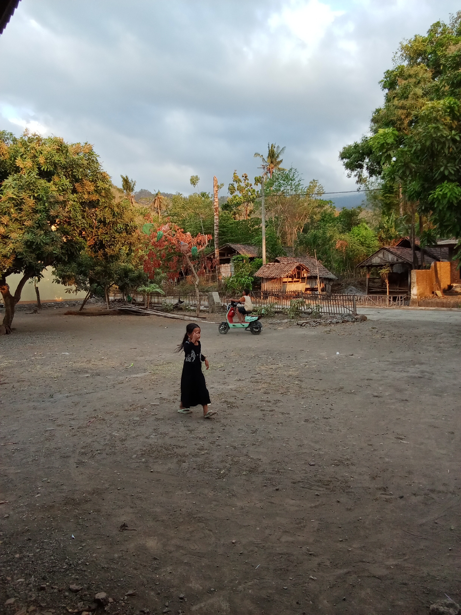 In the city of Selowogo, Indonesia, a young girl with dark hair, wearing a long black dress and sandals, walks across a dusty, open area. Behind her, a person rides a light-colored motor scooter. The background features several rustic, thatched-roof structures, some with open fronts. Dense green trees, including a large tree on the left with a thick trunk, and hills covered in vegetation dominate the landscape under a cloudy sky. The ground is a mixture of dirt and small stones.