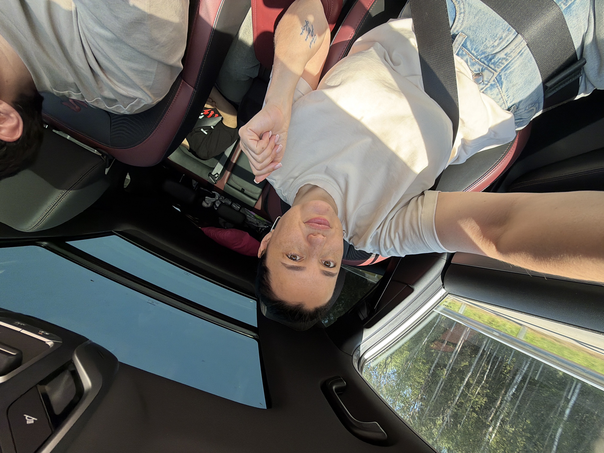 A woman with dark hair and freckles is taking a selfie from the driver's seat of a car. She is wearing a light-colored t-shirt and denim jeans. Her left arm is extended, holding the camera. A tattoo is visible on her forearm. In the passenger seat, a person wearing a black t-shirt is visible. The car has a sunroof, and through the windows, trees and a guardrail are visible, indicating a road passing through a wooded area. The interior of the car features dark-colored seats with red accents and a black ceiling. The scene appears to be set on a sunny day.
